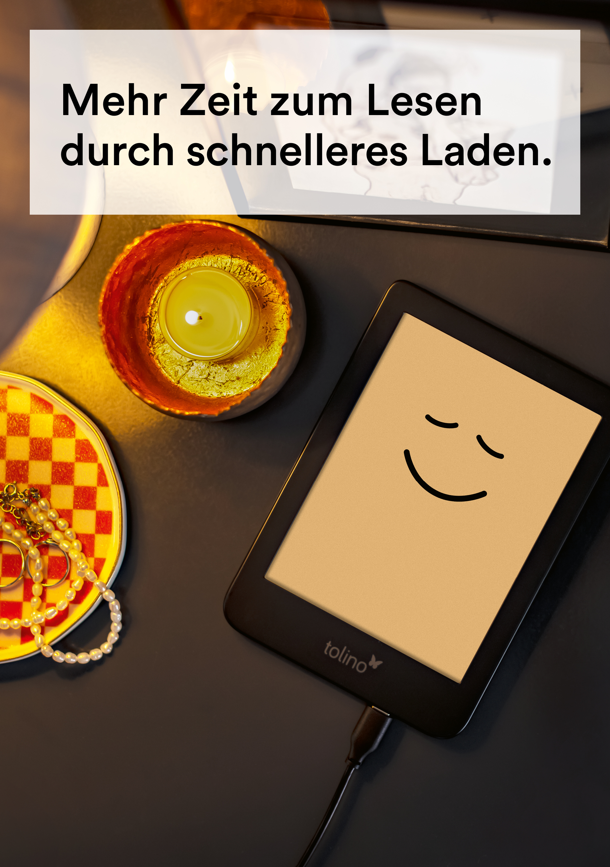 E-Reader mit Smiley, lädt auf Schreibtisch neben Kerze und Schmuck.