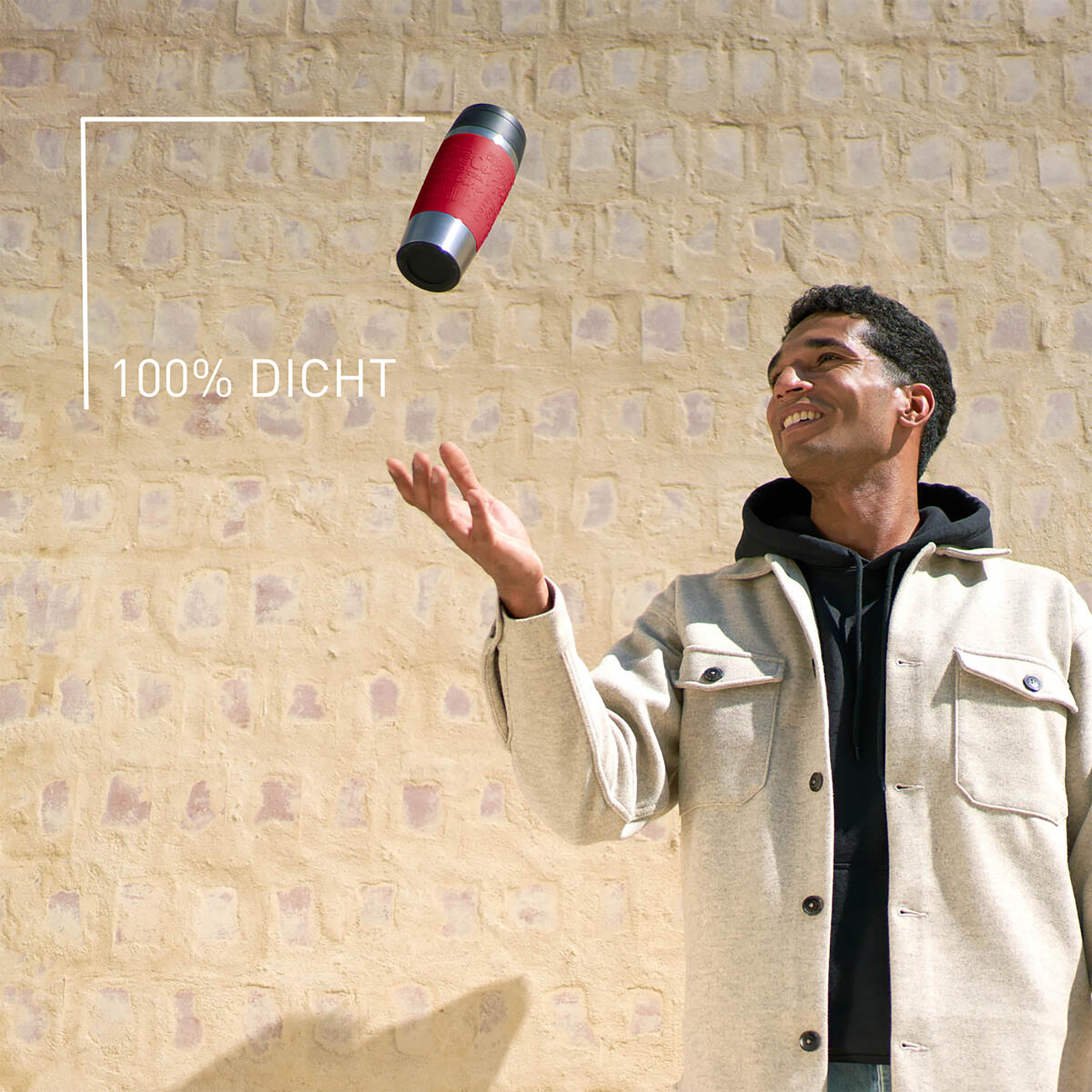 Ein Mann wirft eine rot-silberne Thermosflasche. Der Hintergrund ist eine beige Wand.