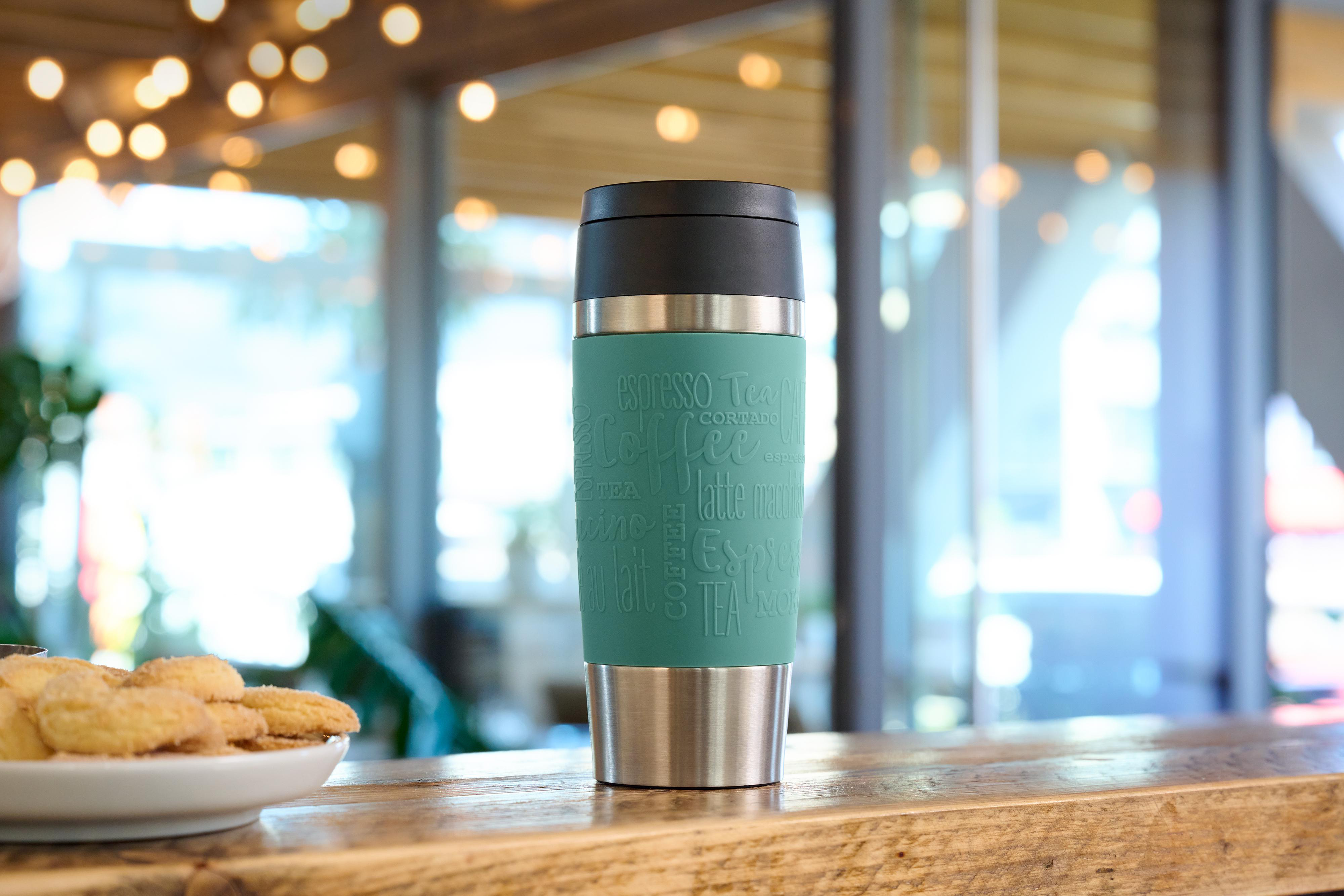 Ein tealfarbener Reisebecher mit Kaffee-Wörtern. Ein Teller mit Keksen im Vordergrund, verschwommener Cafe-Hintergrund.