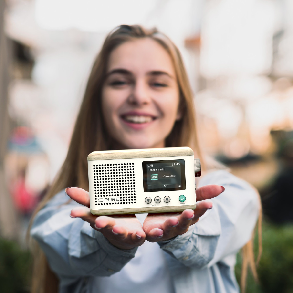 Une femme tient une petite radio. La radio est blanche et possède un écran et des boutons. La femme sourit.