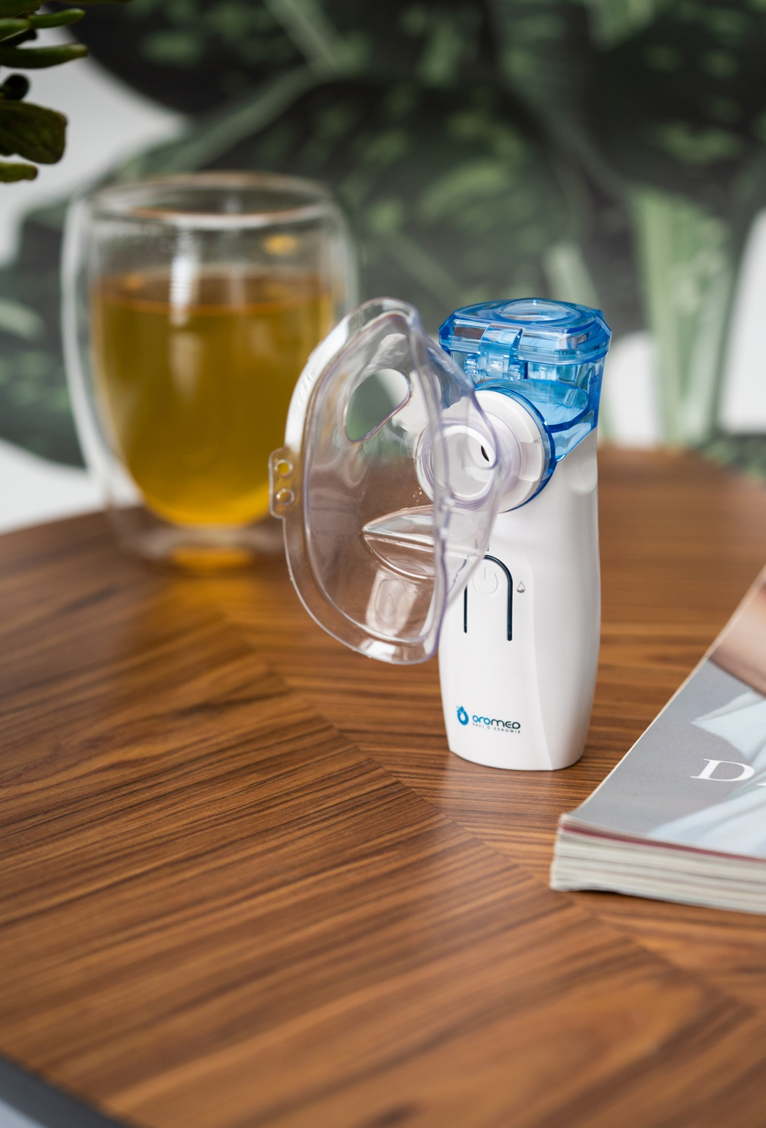 Biało-niebieski nebulizator z maską stoi na drewnianym stole, obok szklanki herbaty i czasopisma.