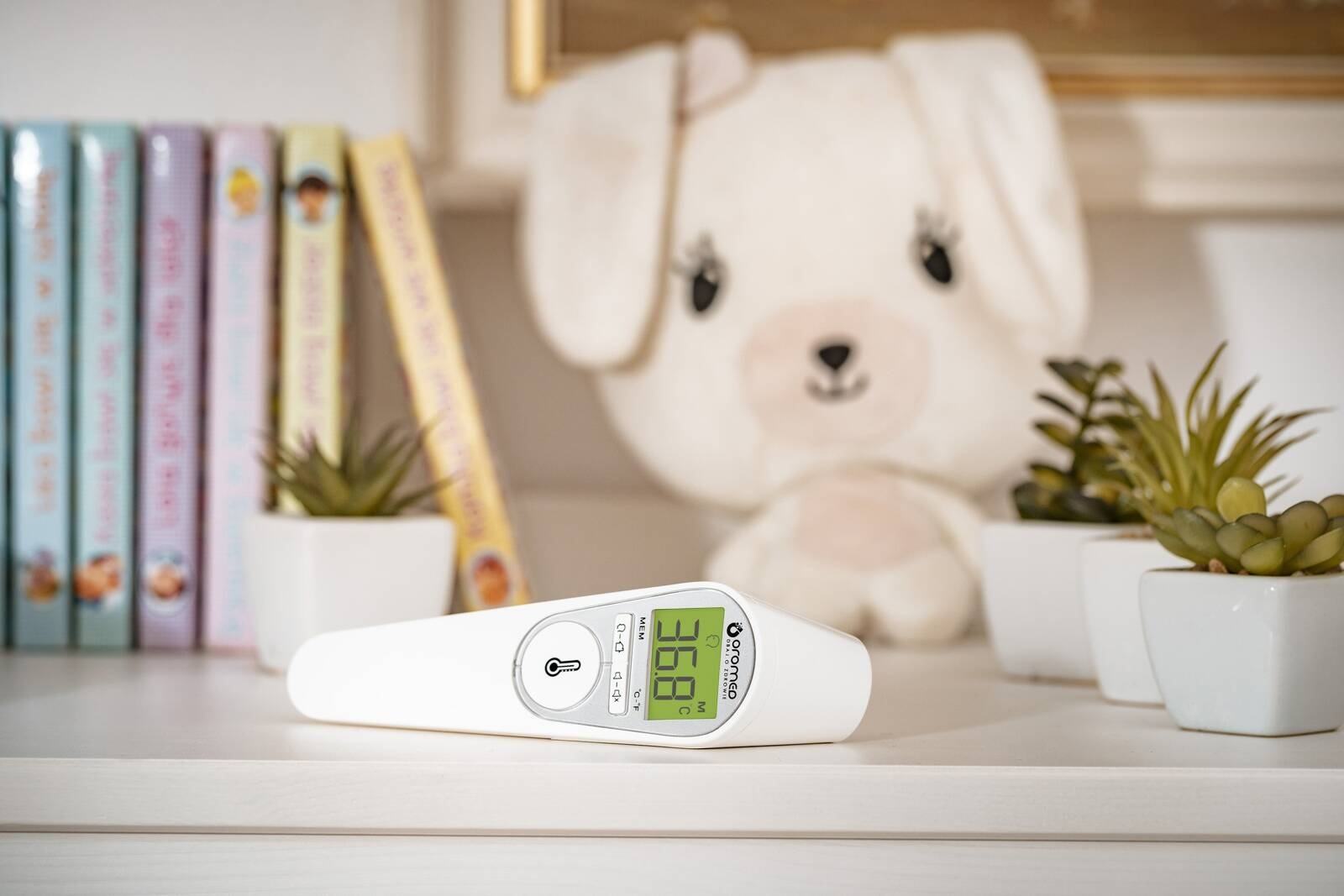 Biały termometr cyfrowy wyświetla 36,8 stopnia. Pluszowy królik i małe rośliny w tle, wraz z książkami.