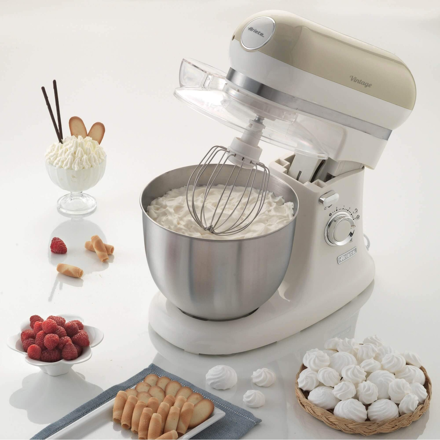 Robot da cucina con ciotola di panna, circondato da meringhe, lamponi e biscotti.