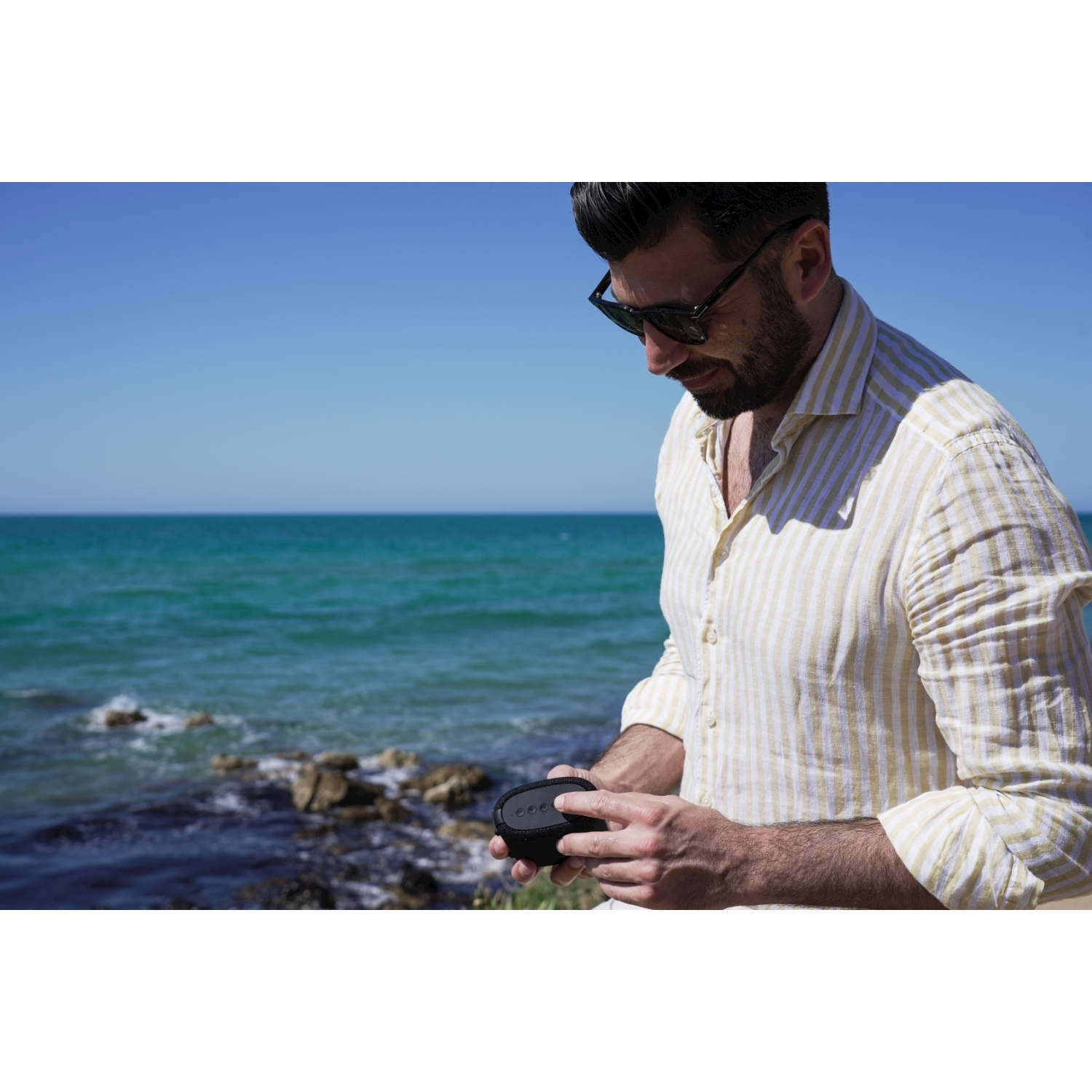 Un uomo con occhiali da sole e una camicia a righe guarda un piccolo altoparlante nero.