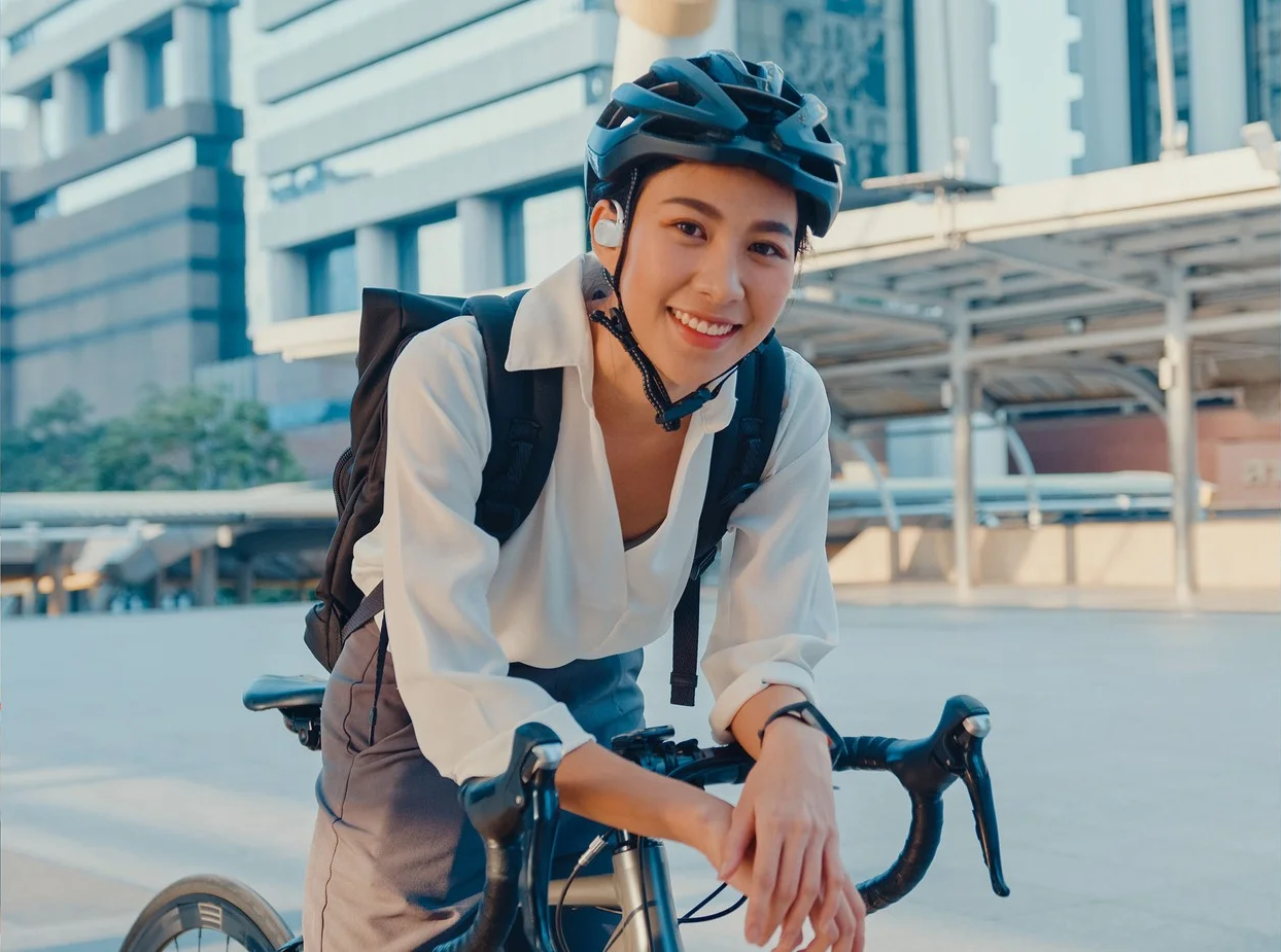 Frau mit Helm, Fahrradfahren, lächelnd, mit Gebäuden im Hintergrund.