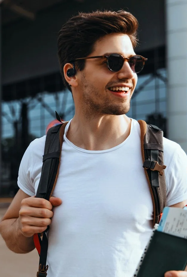 Ein Mann mit Sonnenbrille und Ohrhörern lächelt und hält eine Bordkarte. Er hat einen Rucksack auf.
