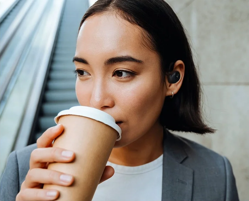 Frau mit Ohrhörern trinkt aus einer Kaffeetasse in der Nähe einer Rolltreppe.