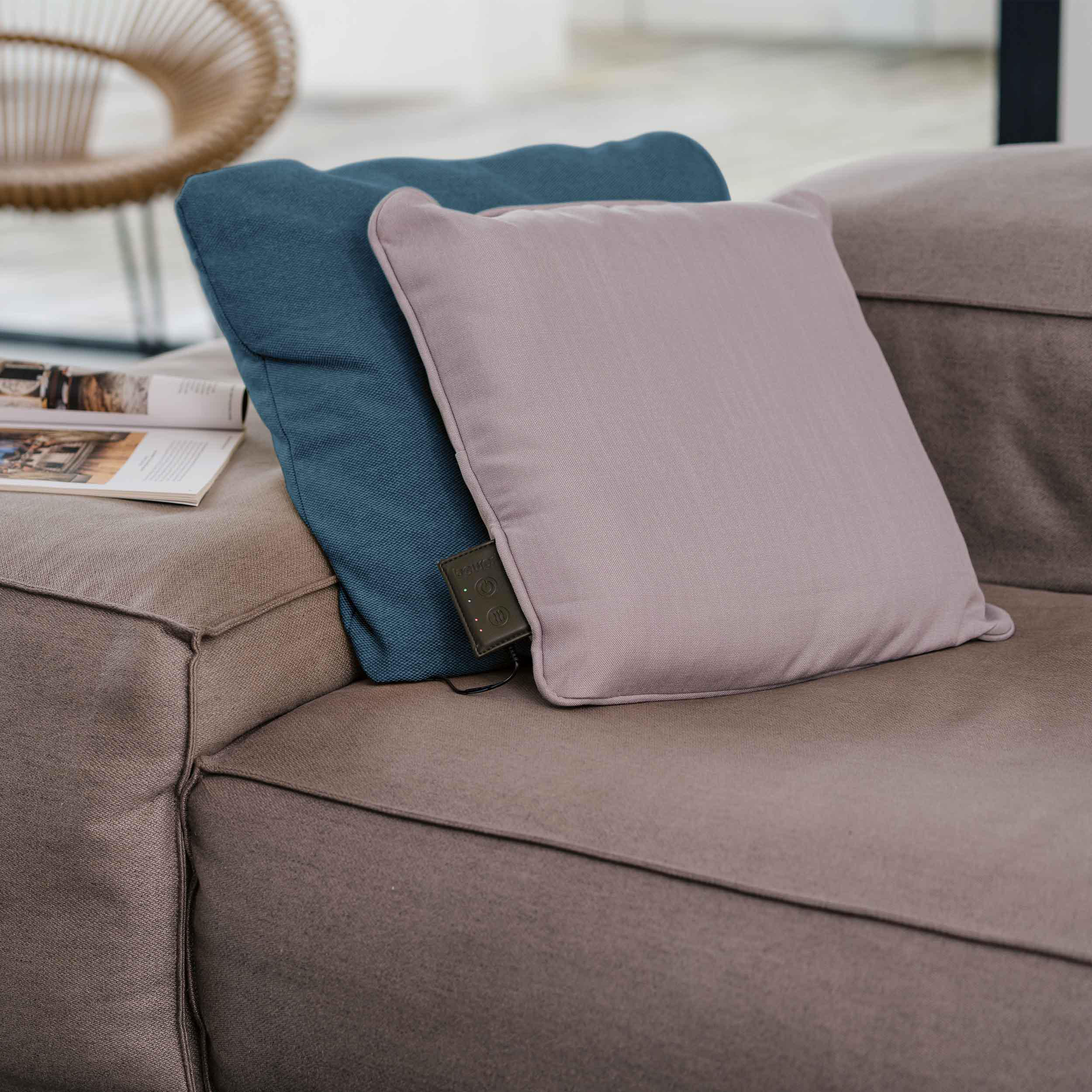 Zwei quadratische Kissen, eines blau, eines rosa, auf einem braunen Sofa. Ein Buch liegt daneben.