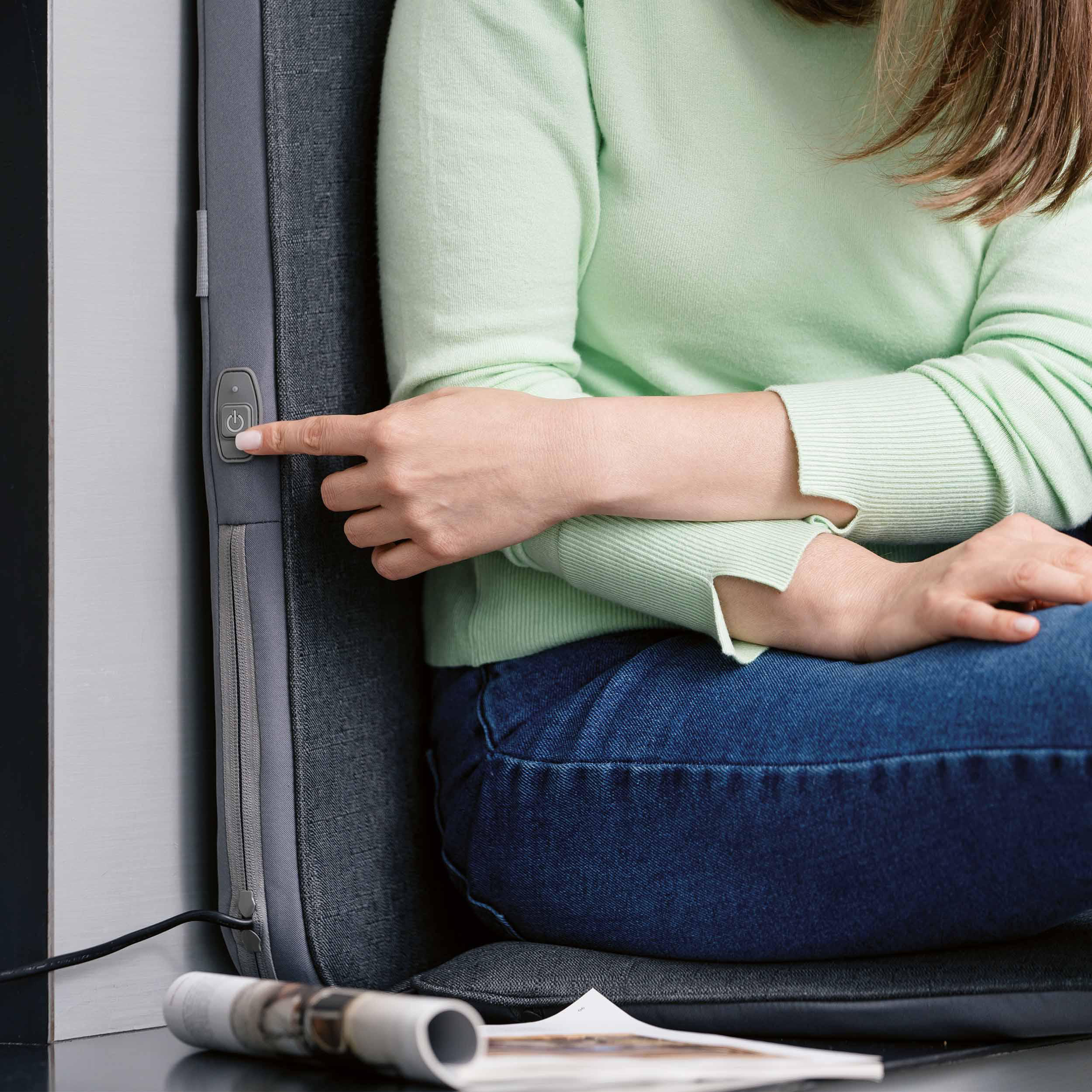 Eine Frau drückt einen Knopf auf einem Stuhl. Sie trägt Jeans und einen grünen Pullover. Eine Zeitschrift liegt.