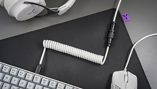 Czarna mysz, klawiatura i biały kabel spiralny na czarnej podkładce. Białe słuchawki w tle.