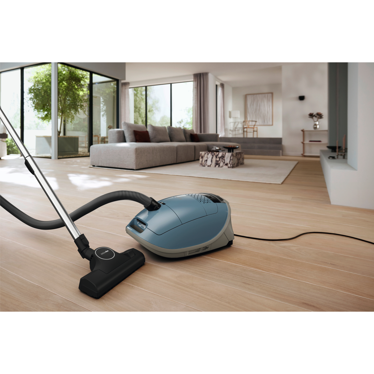 A blue vacuum cleaner with a hose and head on wooden floor. Room with sofa and a tree.