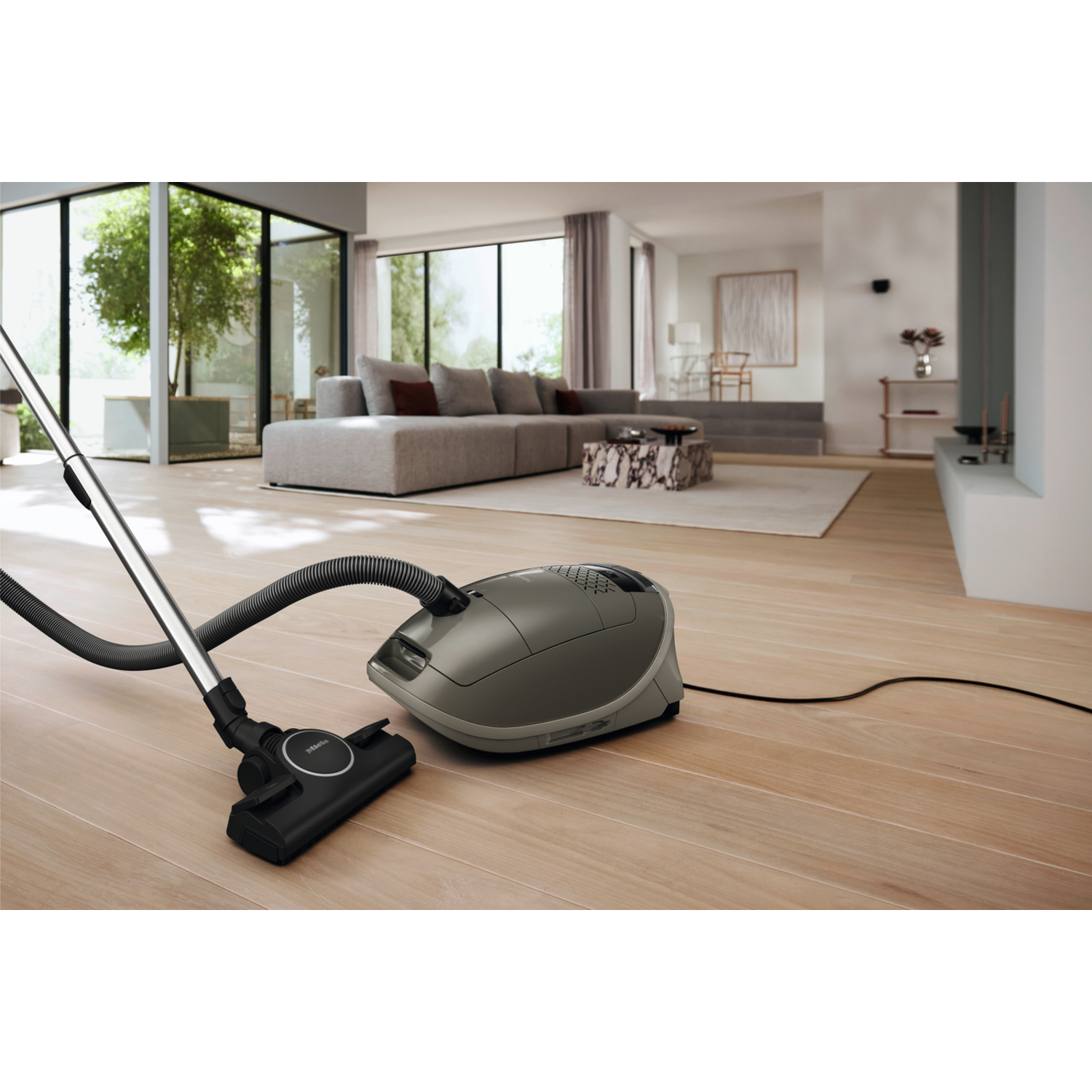 Vacuum cleaner on wooden floor, with a sofa and windows in the background. 