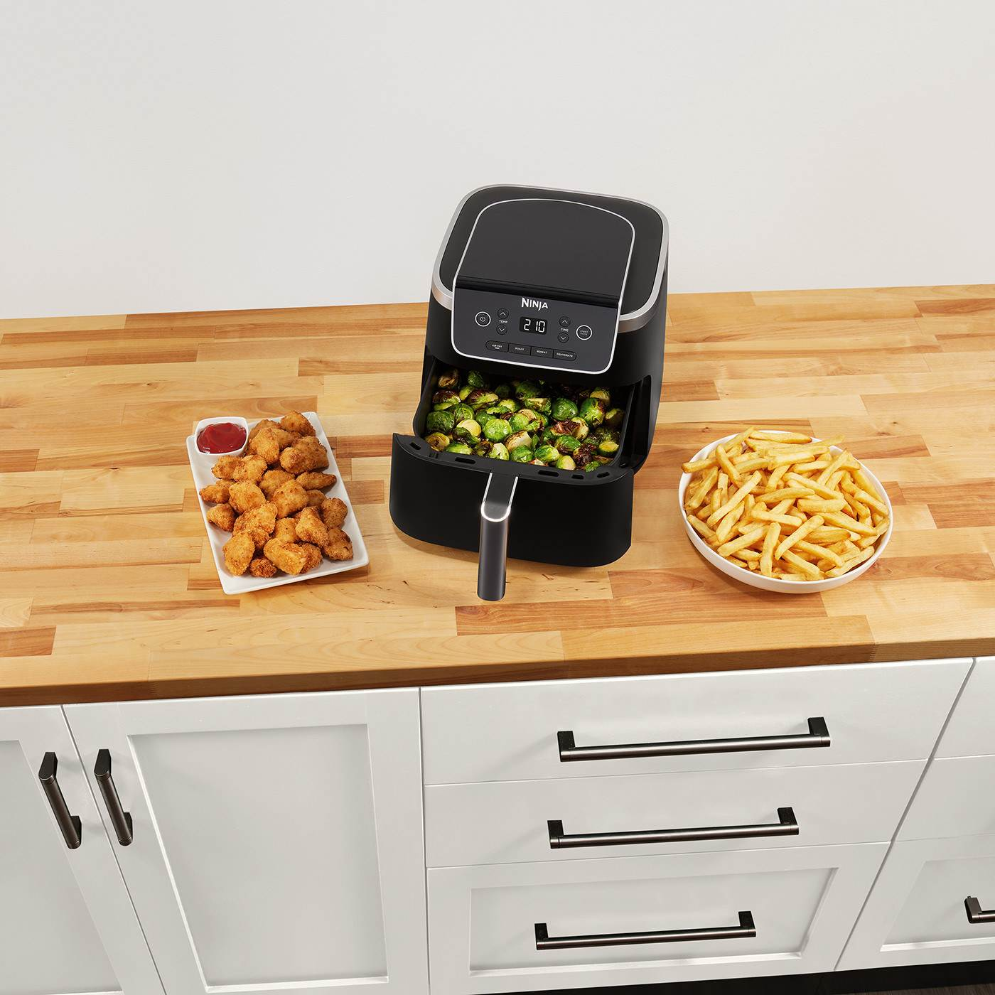 Friggitrice ad aria con cibo all'interno, accanto a un piatto di crocchette e patatine fritte.