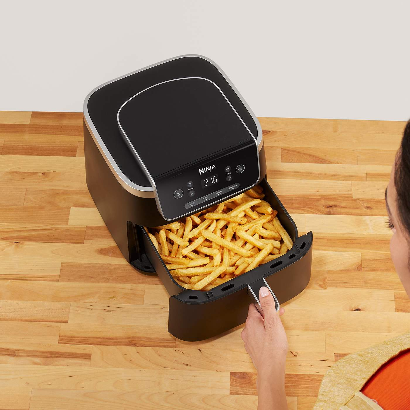 Friggitrice ad aria Ninja nera con patatine fritte cotte nel suo cestello, su una superficie di legno, tenuta da una persona.
