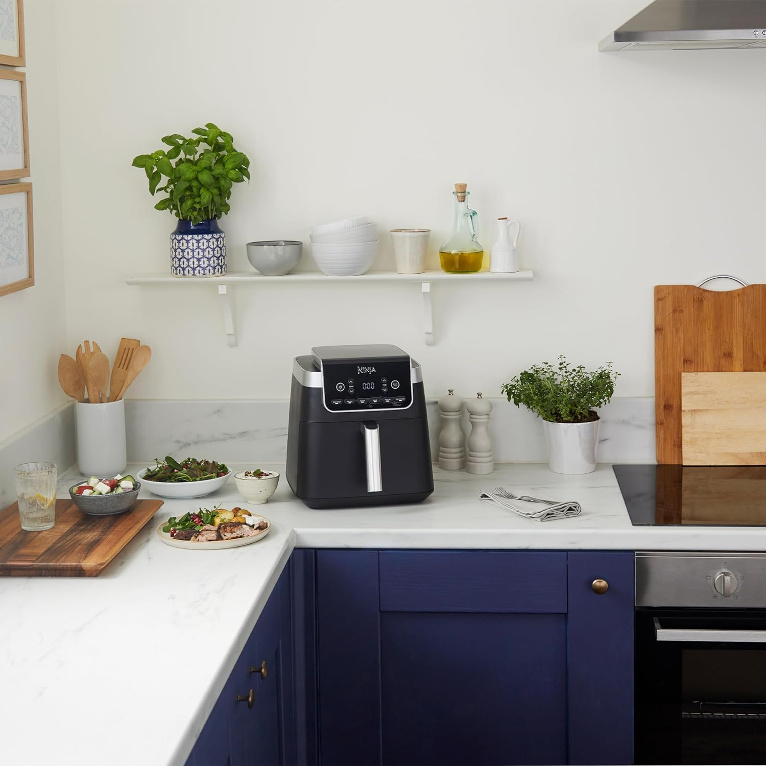 Cucina con friggitrice ad aria, piante, cibo e armadietti blu. Mensole e piano di lavoro bianchi.