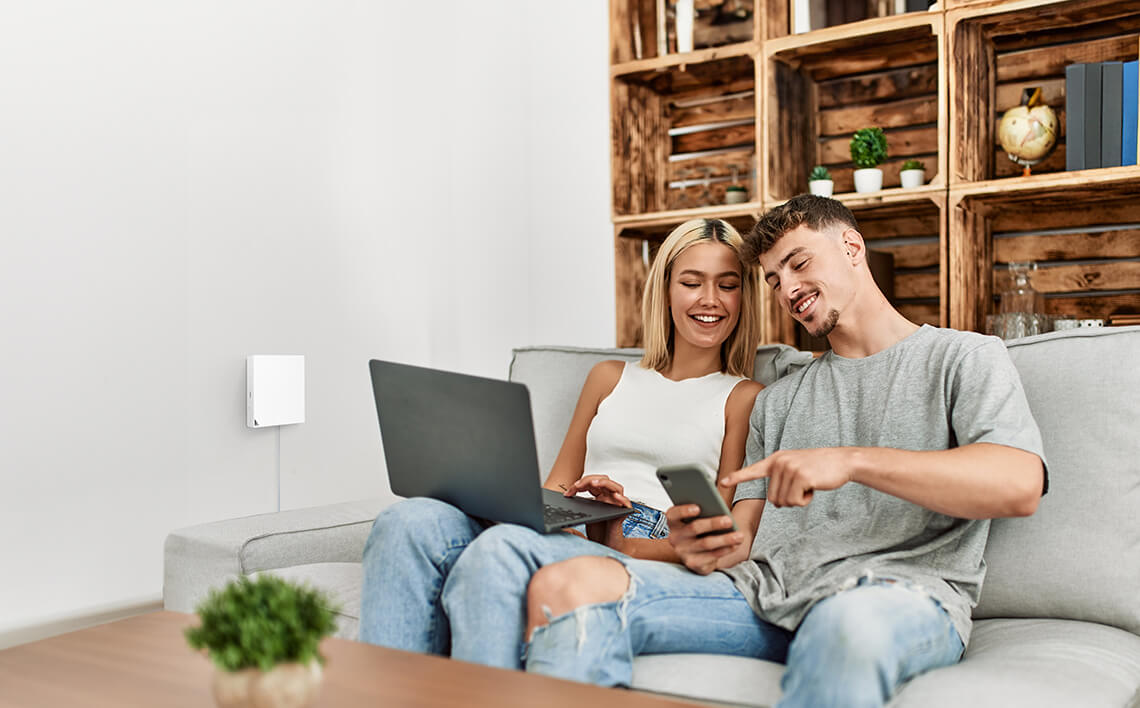 Un couple assis sur un canapé regarde un téléphone et un ordinateur portable. Arrière-plan: étagères en bois et mur.