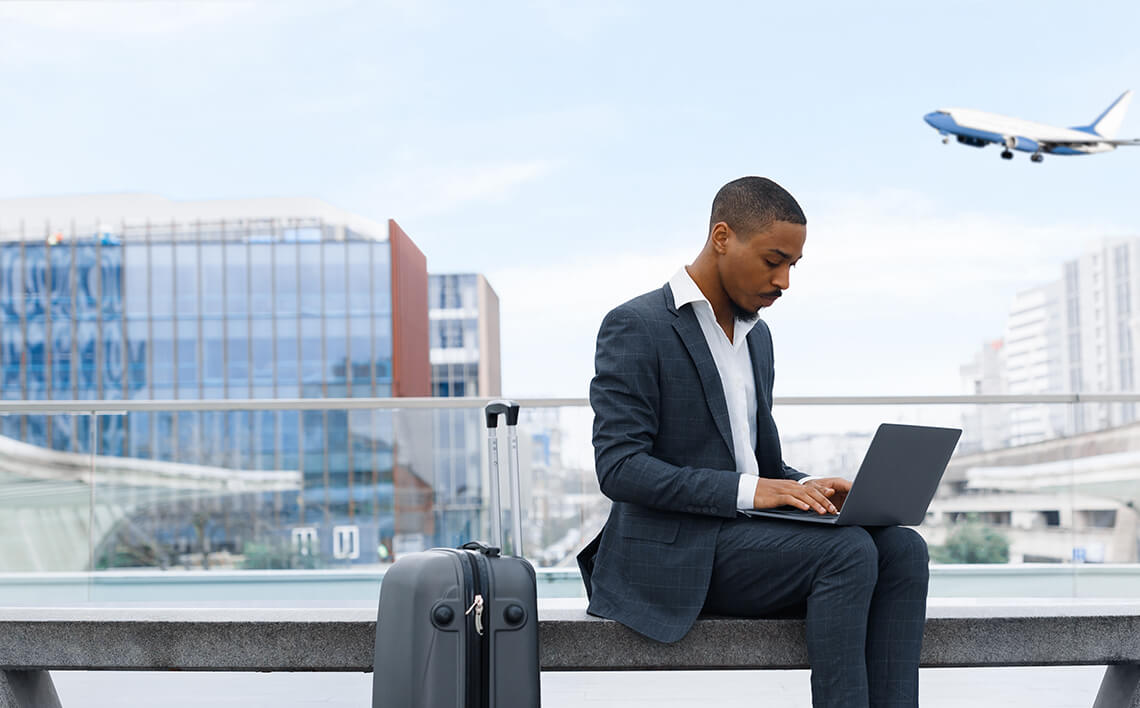 Homme avec ordinateur portable sur un banc, avion en arrière-plan, valise à proximité.