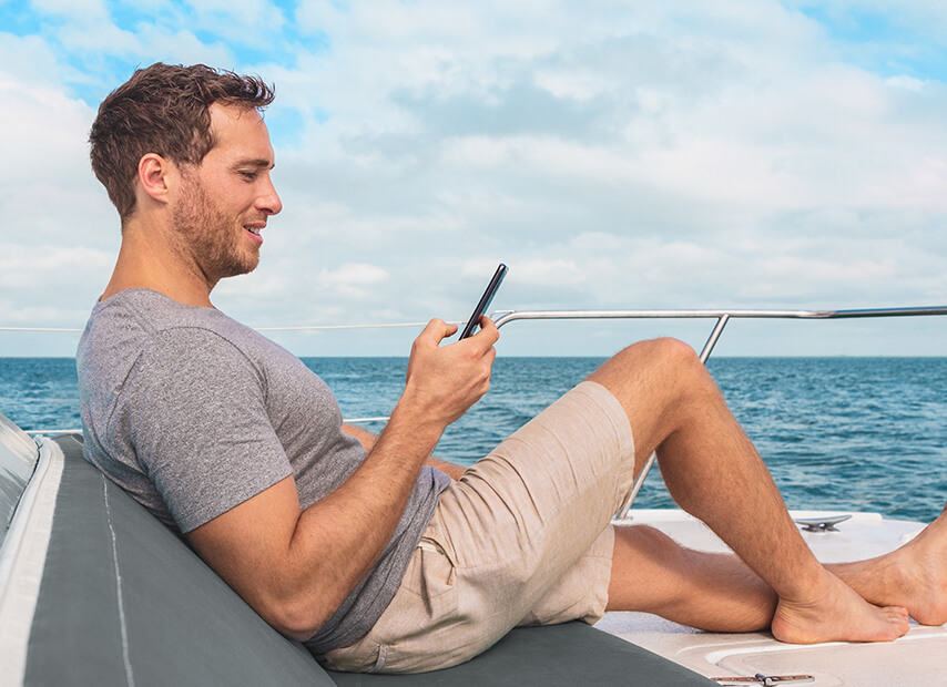 Un homme sur un bateau, assis et regardant son téléphone. Il porte un short et un t-shirt, avec de l'eau en arrière-plan.