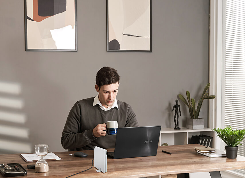 Un homme travaille sur un ordinateur portable sur un bureau en bois, tenant une tasse. Il y a une lampe.