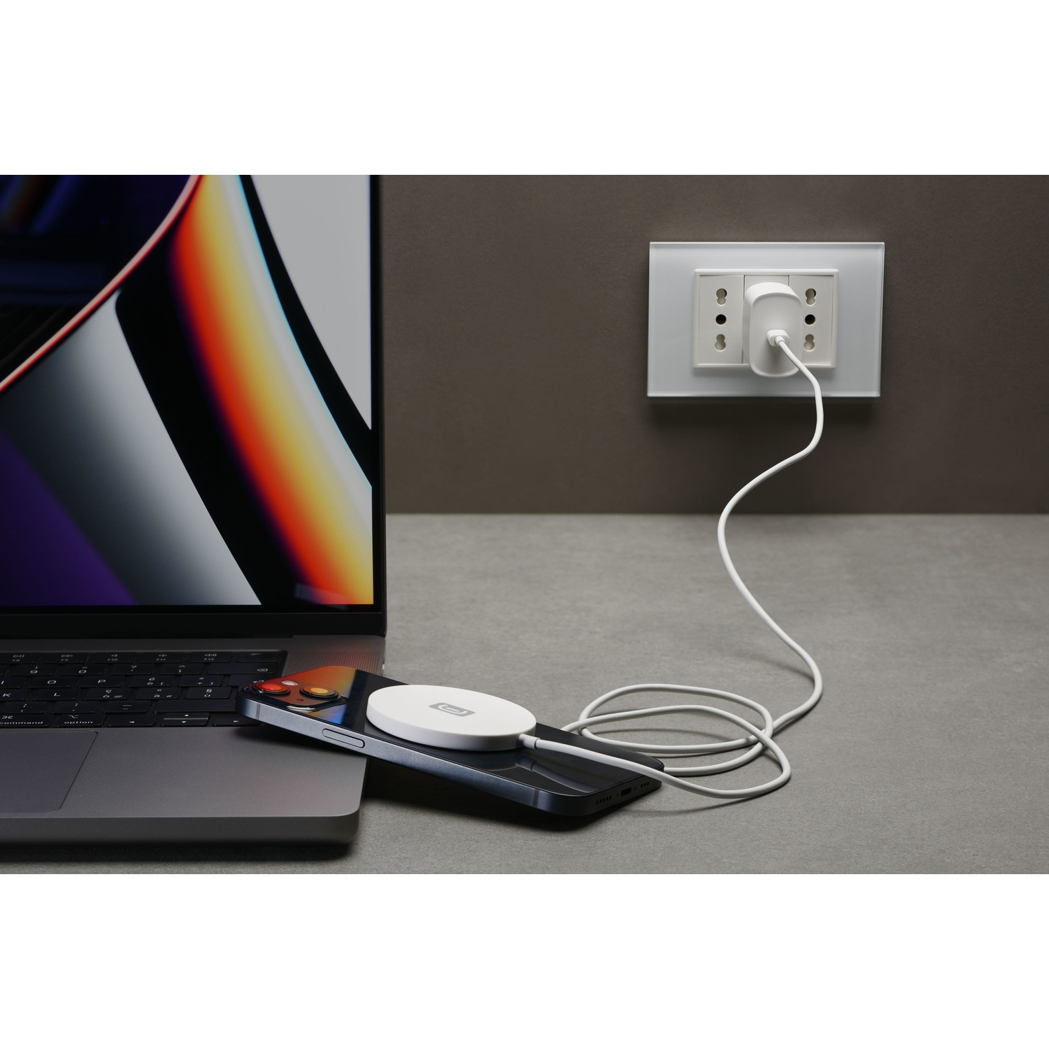 Laptop collegato a un caricatore wireless bianco su un telefono, collegato a una presa a muro su una superficie grigia.