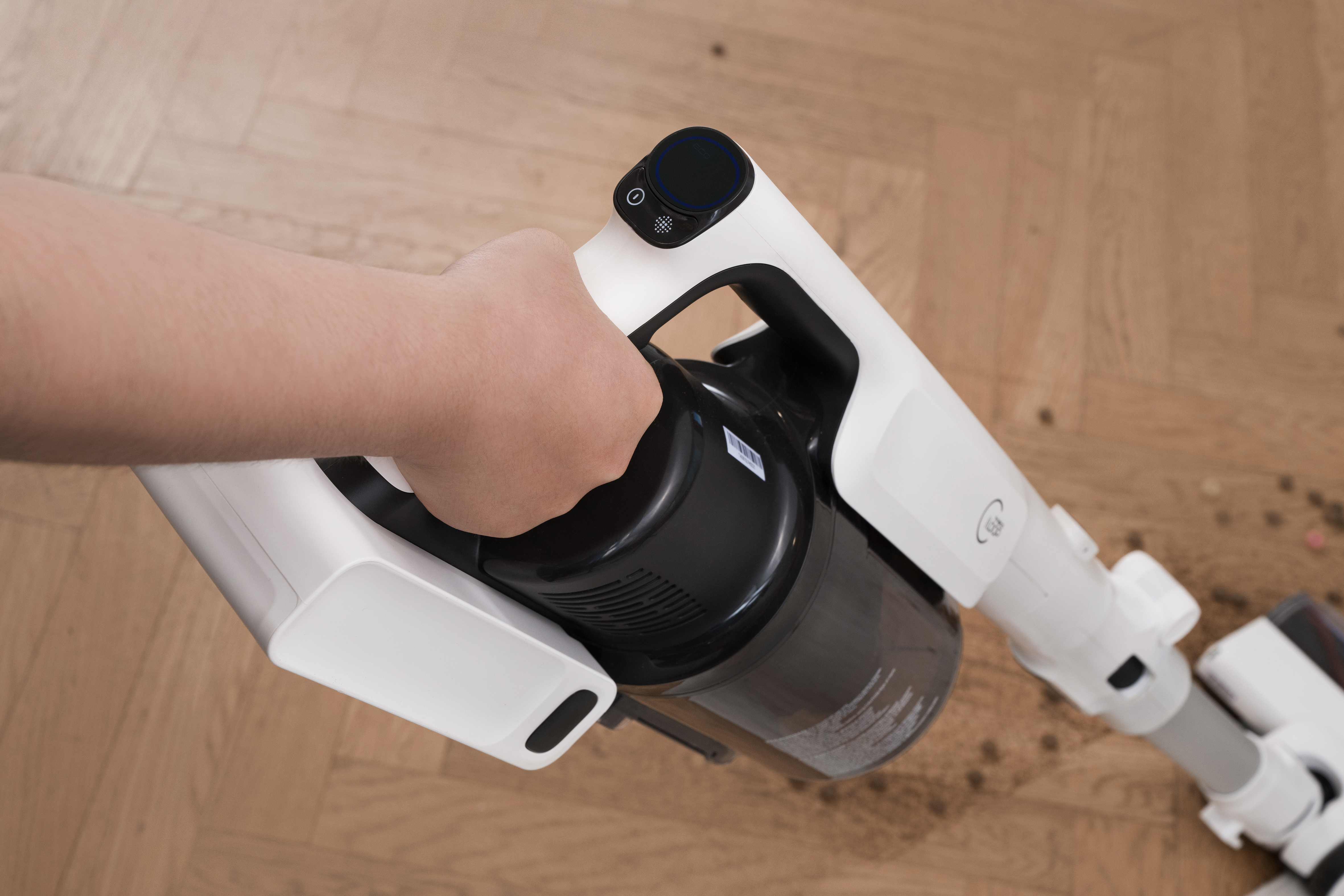Eine Hand hält einen weiß-schwarzen Staubsauger auf einem Holzboden, mit herumliegenden Trümmern.