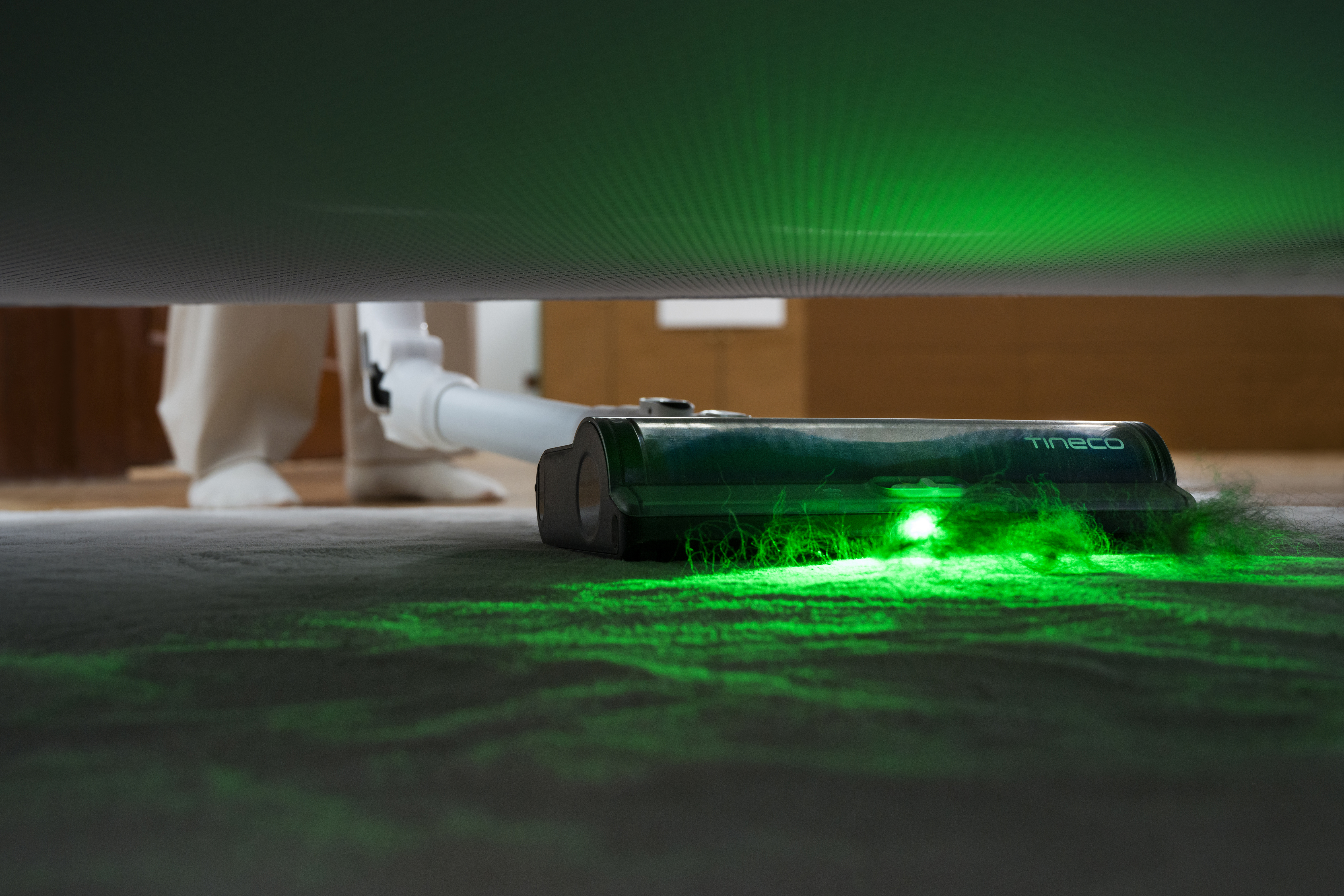 Staubsaugerkopf mit grünem Laserlicht, der einen Teppich reinigt. Die Beine einer Person sind sichtbar.