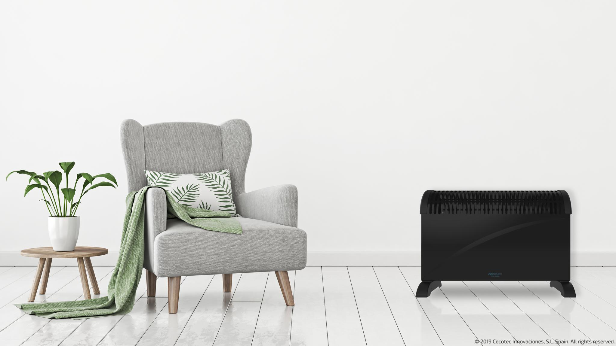 Calefactor negro cerca de una silla gris, manta verde. Paredes blancas y suelo de madera.