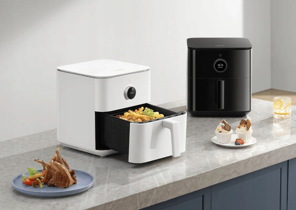 Dwie frytkownice powietrzne, jedna biała, druga czarna, z jedzeniem i dodatkami na blacie.