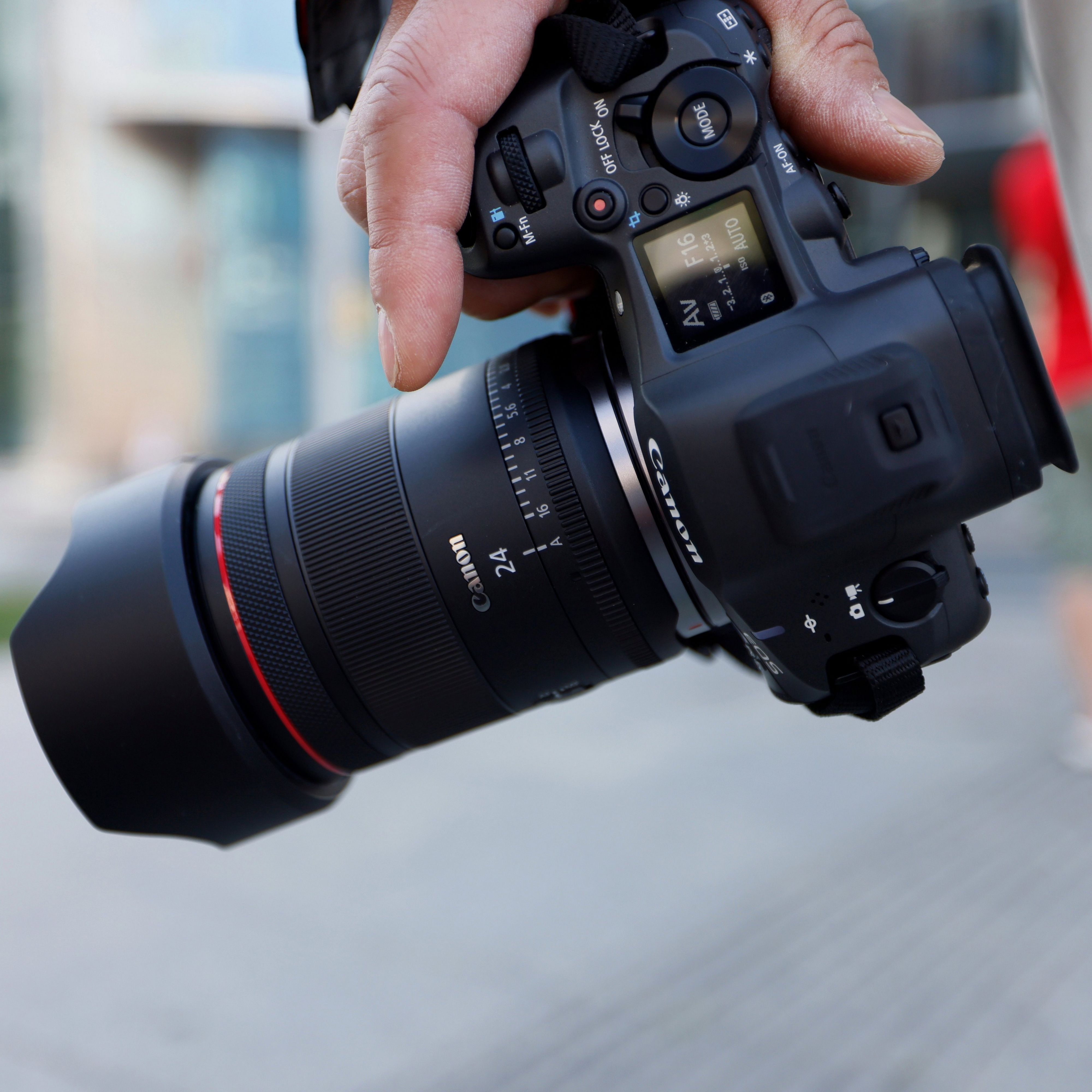 Une main tient un appareil photo Canon avec objectif noir à anneau rouge. Arrière-plan flou.