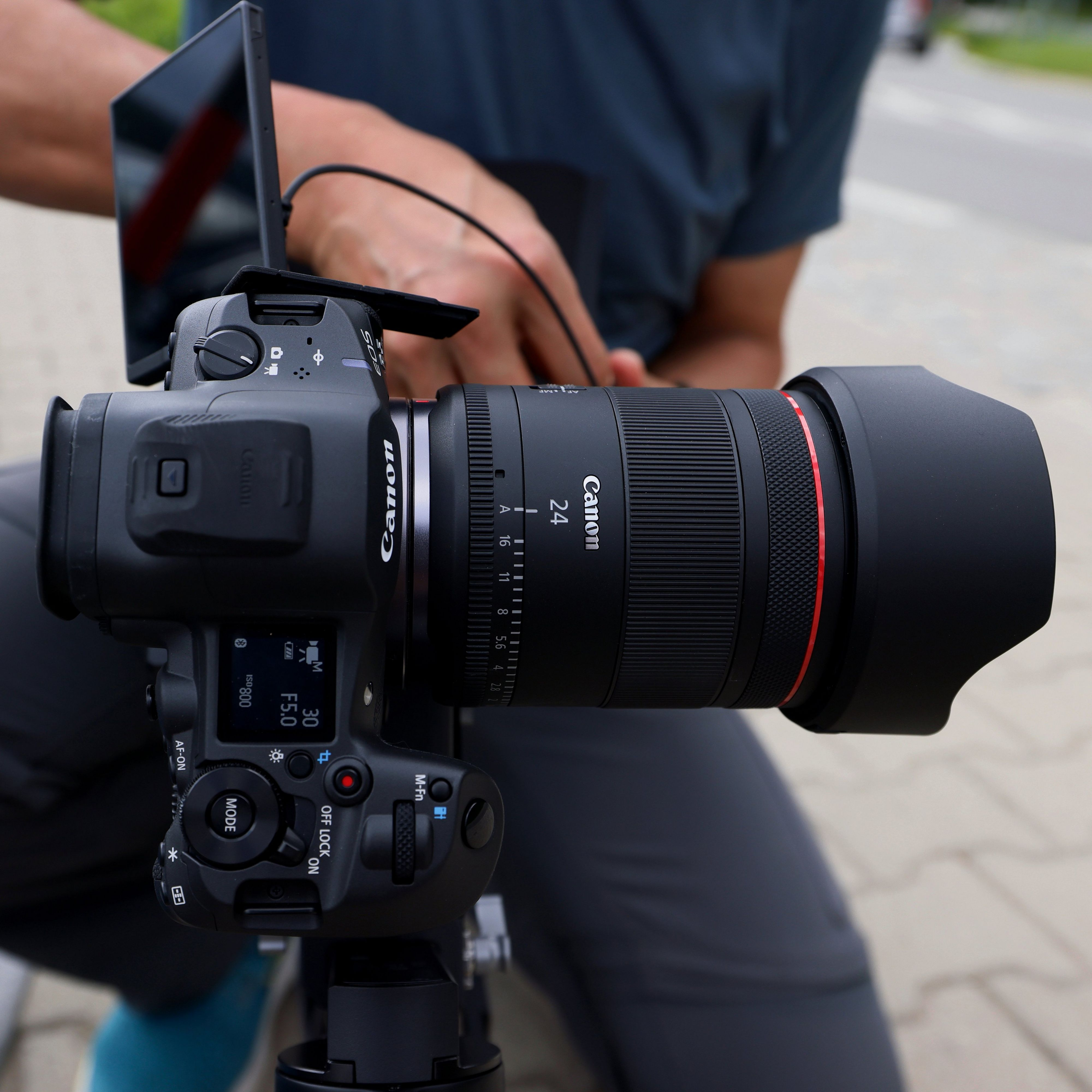 Un appareil photo Canon avec un objectif est tenu par une personne. L'objectif est noir, anneau rouge.