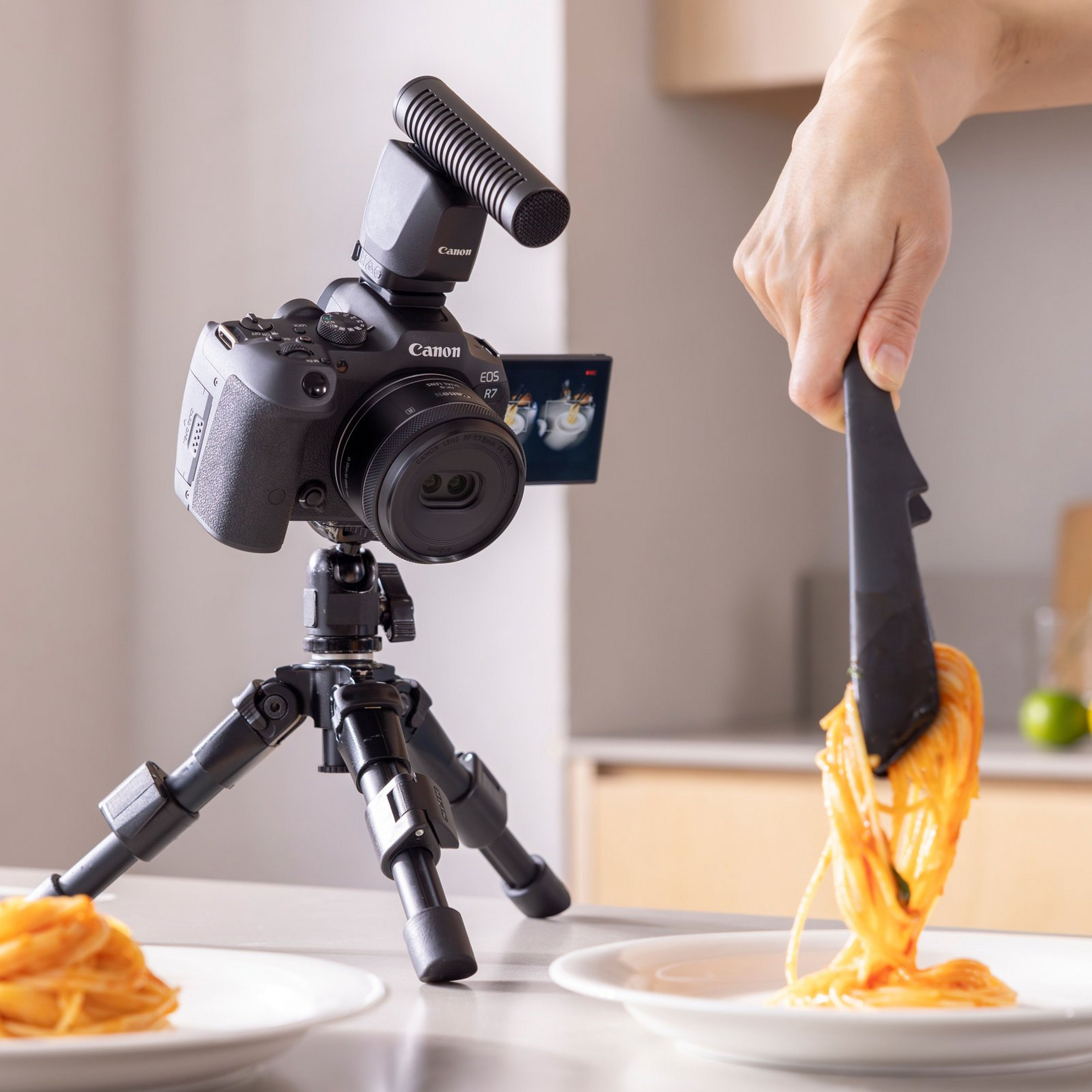 Une caméra sur un trépied avec un microphone et une main utilisant des pinces pour servir des spaghettis.