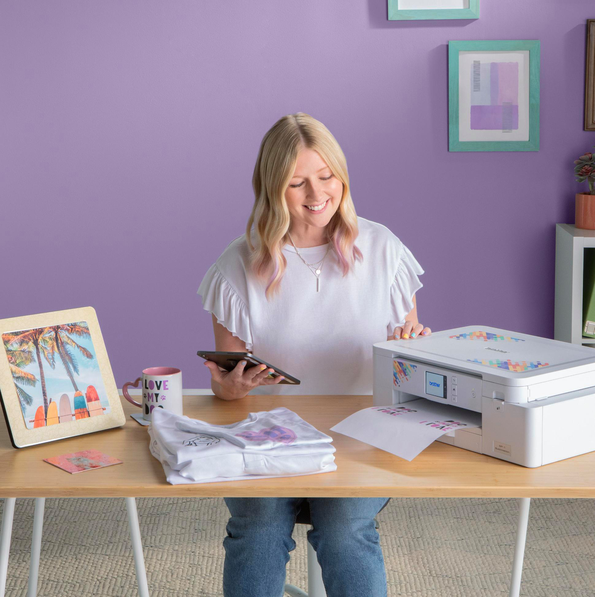 Frau lächelt am Drucker, hält ein Tablet. T-Shirt auf dem Tisch mit Bilderrahmen und Tasse.