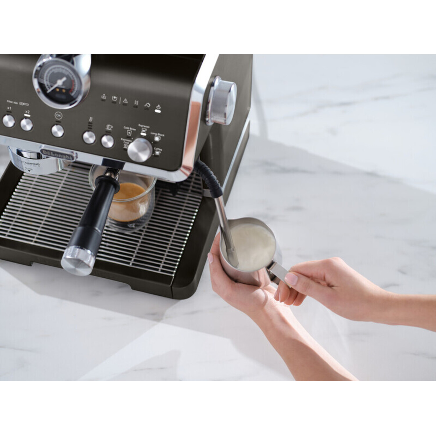 A person using an espresso machine. They are steaming milk into a metal pitcher, with coffee in a glass.