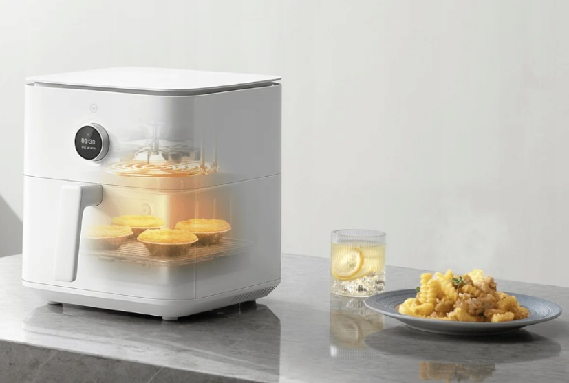 Biała frytkownica powietrzna z jedzeniem w środku, szklanka wody z cytryną i talerz smażonego jedzenia.