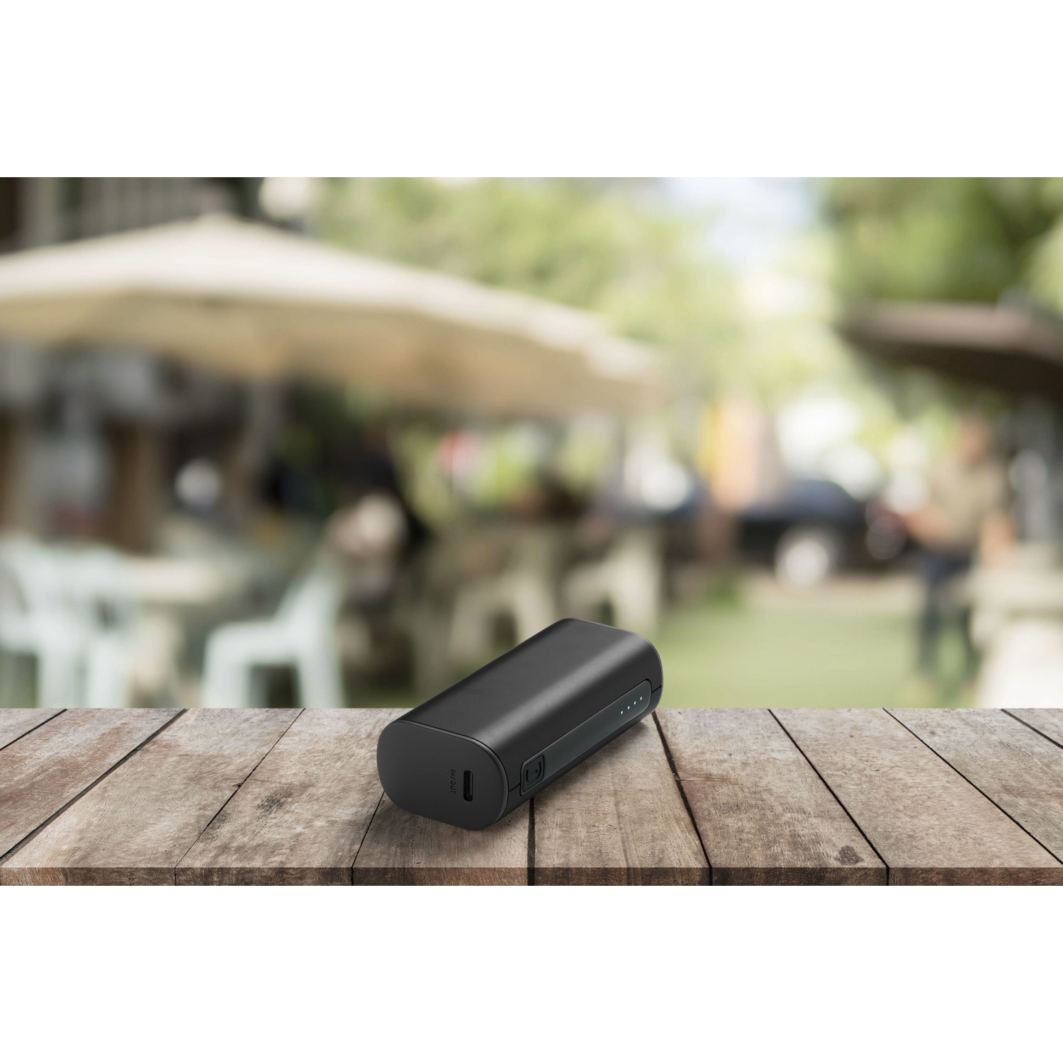 Un power bank nero su un tavolo di legno. Lo sfondo è una scena sfocata all'aperto con persone.