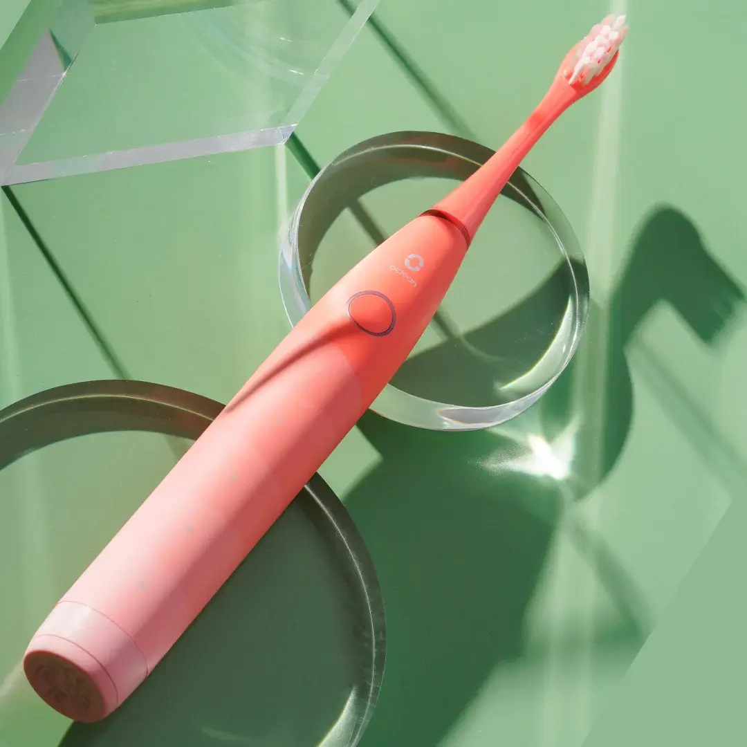 Różowa elektryczna szczoteczka do zębów na przezroczystym, okrągłym stojaku. Jasnozielone tło.
