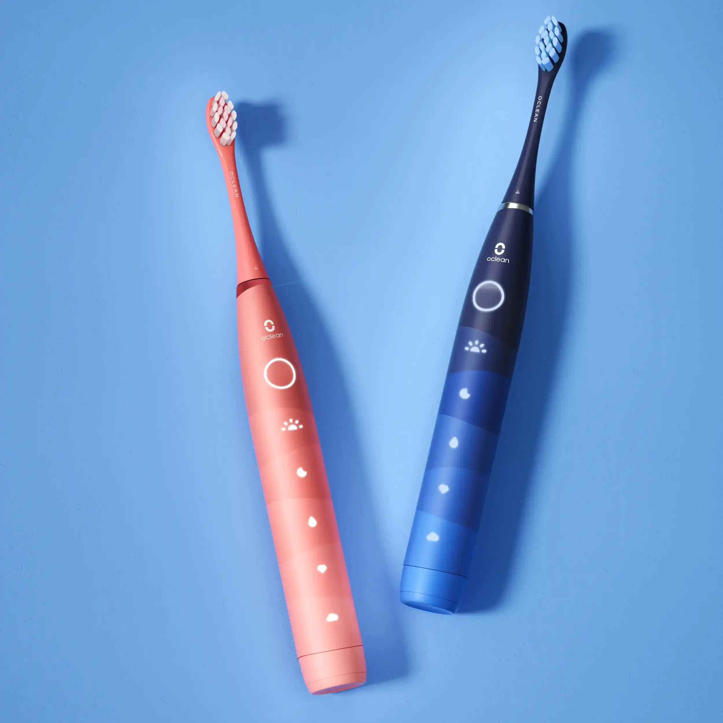 Dwie elektryczne szczoteczki do zębów, jedna koralowa i jedna niebieska, na niebieskim tle. Mają ten sam design.