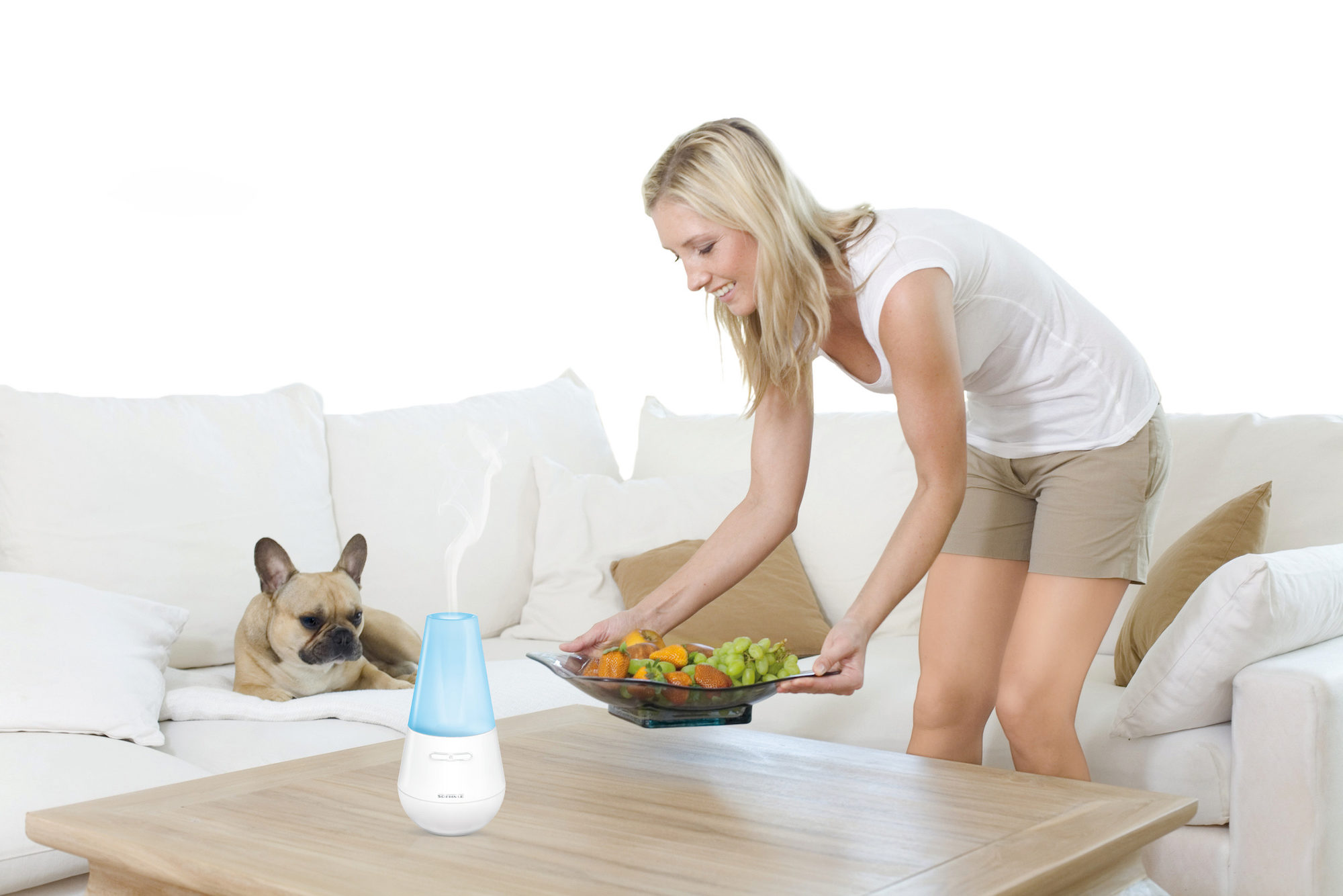 Kobieta stawia miskę z owocami, dyfuzor i pies na kanapie, jasne otoczenie.