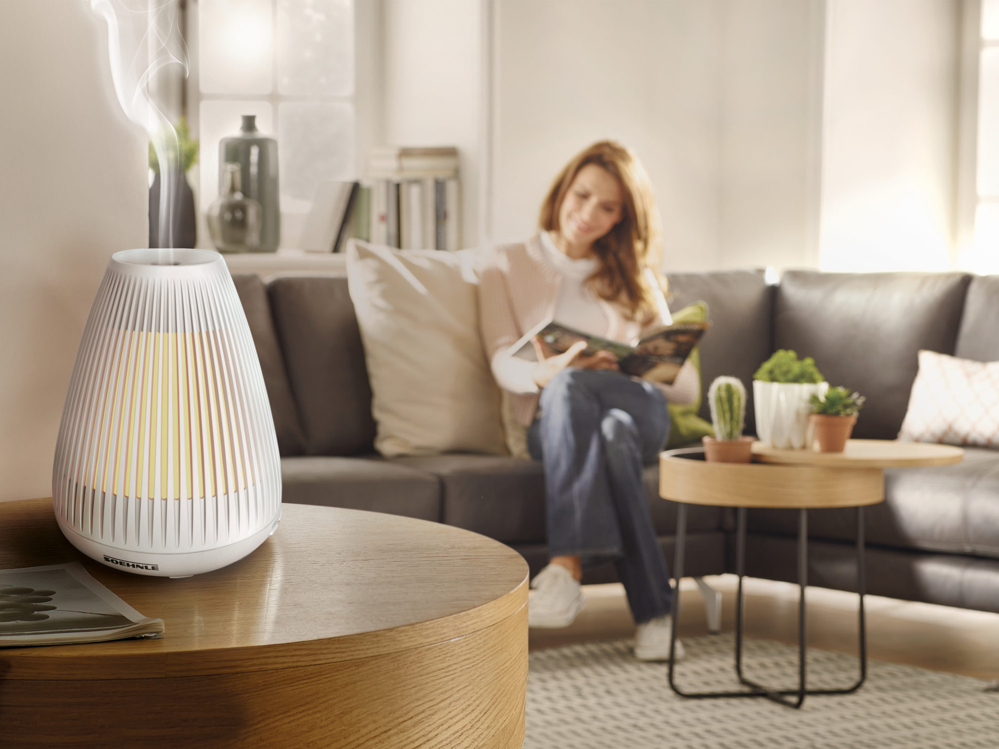 Biały dyfuzor olejków eterycznych z kobietą czytającą książkę w tle.