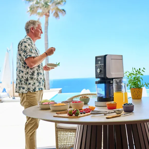 Adam açık havada yemek hazırlıyor. Masada Ninja blender, meyveler ve mavi gökyüzü ile meyve suyu var.