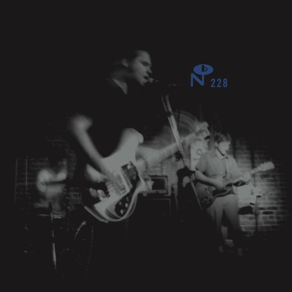 Monochrome Bild einer live auftretenden Band. Die Band spielt auf dunkler Bühne, mit Backsteinmauer.
