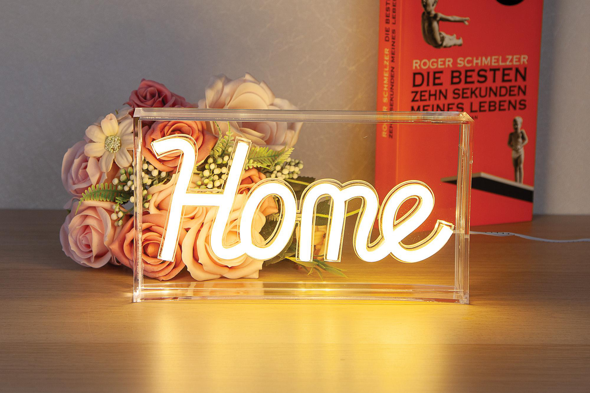 Beleuchtetes "Home"-Schild. Blumen und ein Buch sind sichtbar. Schild auf Tisch.