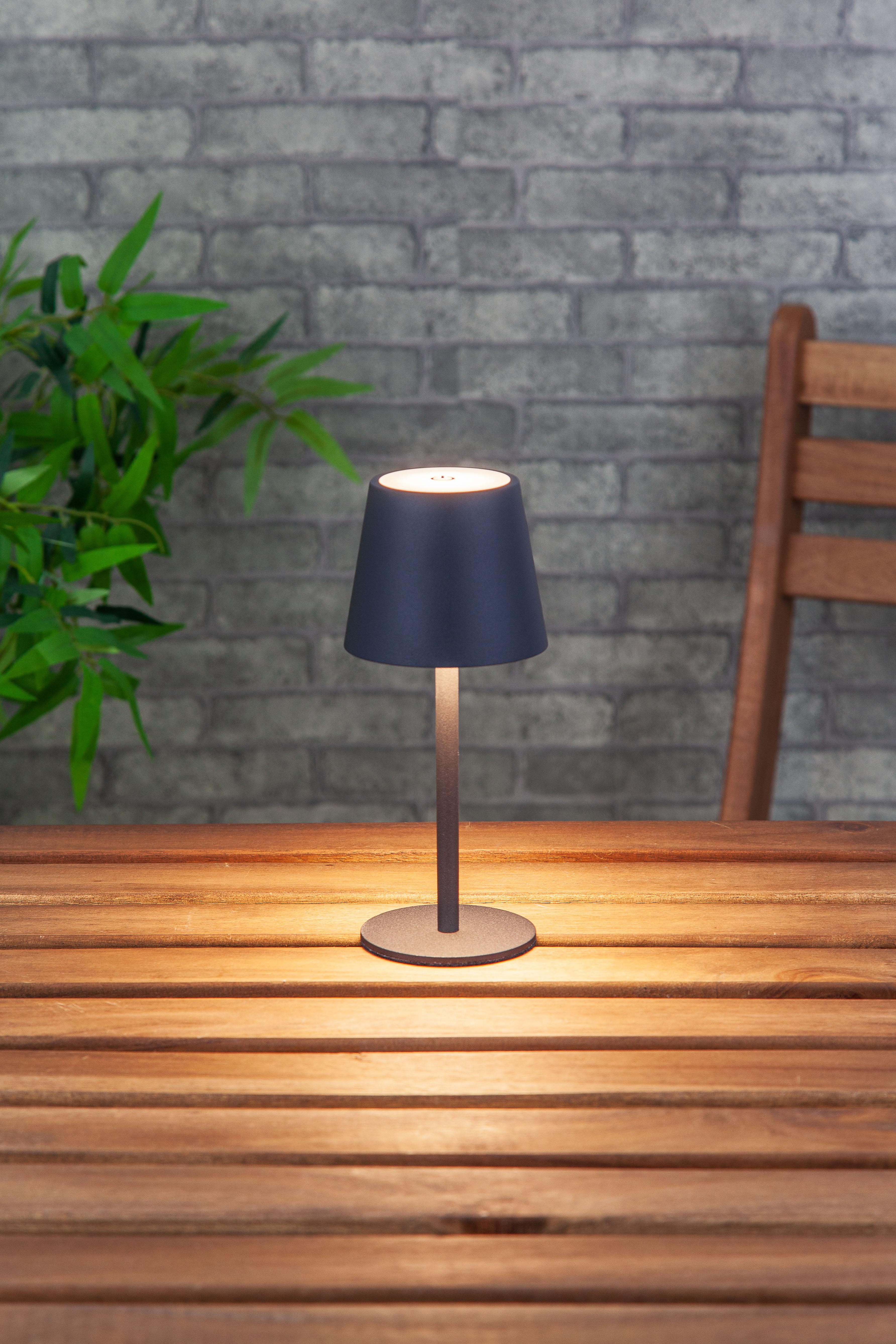 Tischlampe auf einem Holztisch mit Backsteinmauer im Hintergrund. Die Lampe leuchtet.