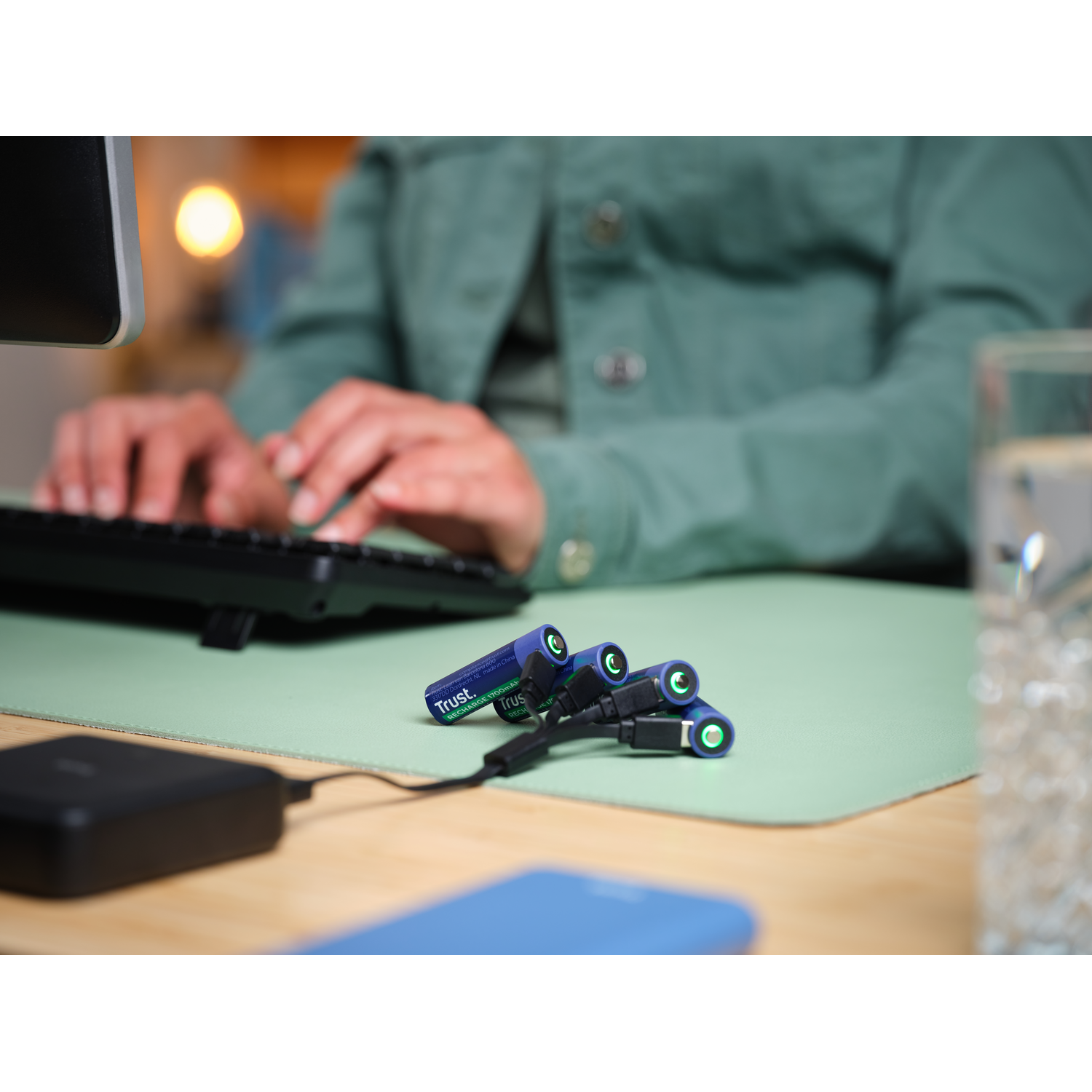 A person is working at a desk, with batteries charging. Green desk mat. Black keyboard and monitor.