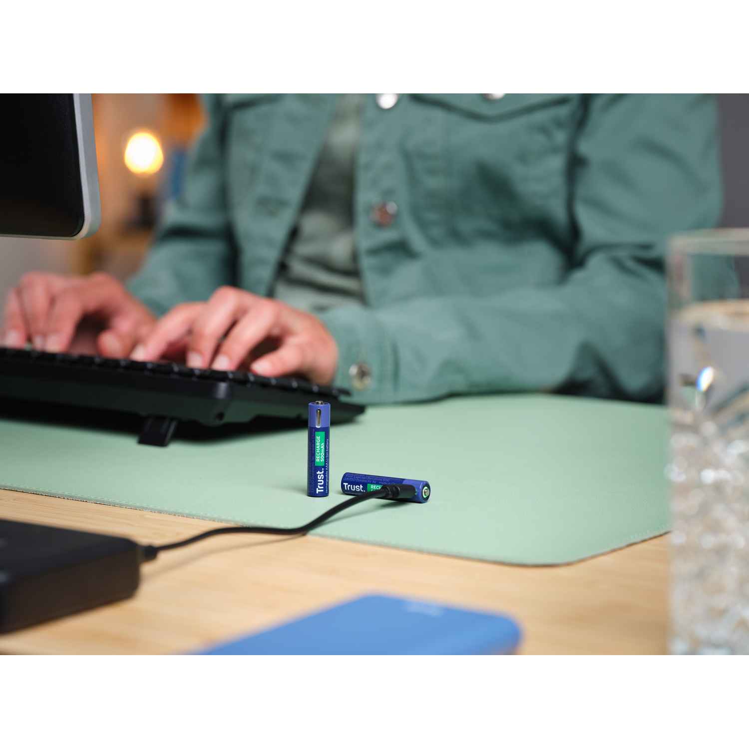 Two blue and green Trust batteries with a keyboard and a glass of water.