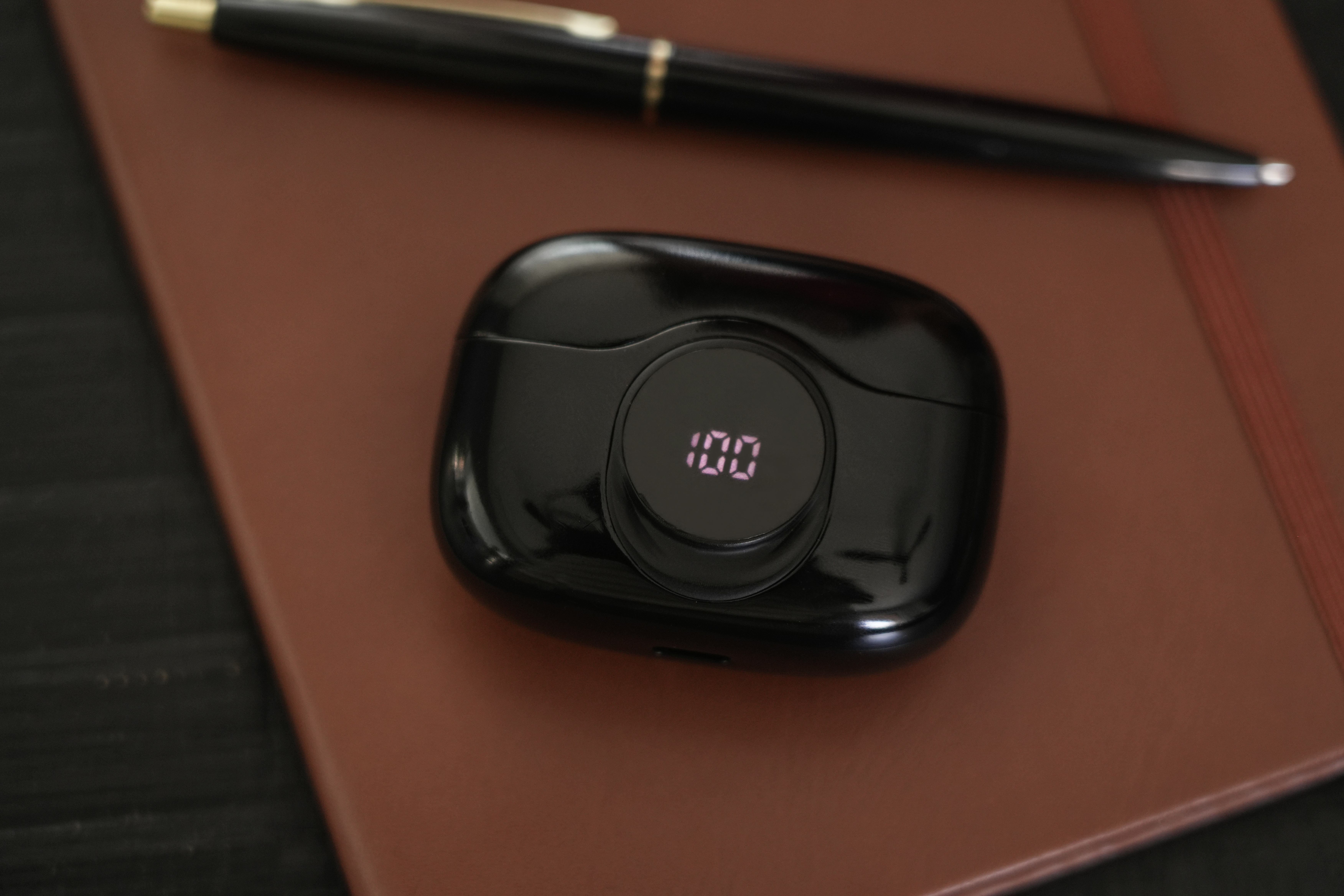 Schwarzes Ladecase mit Digitalanzeige '100' neben einem Stift auf einem braunen Notizblock.