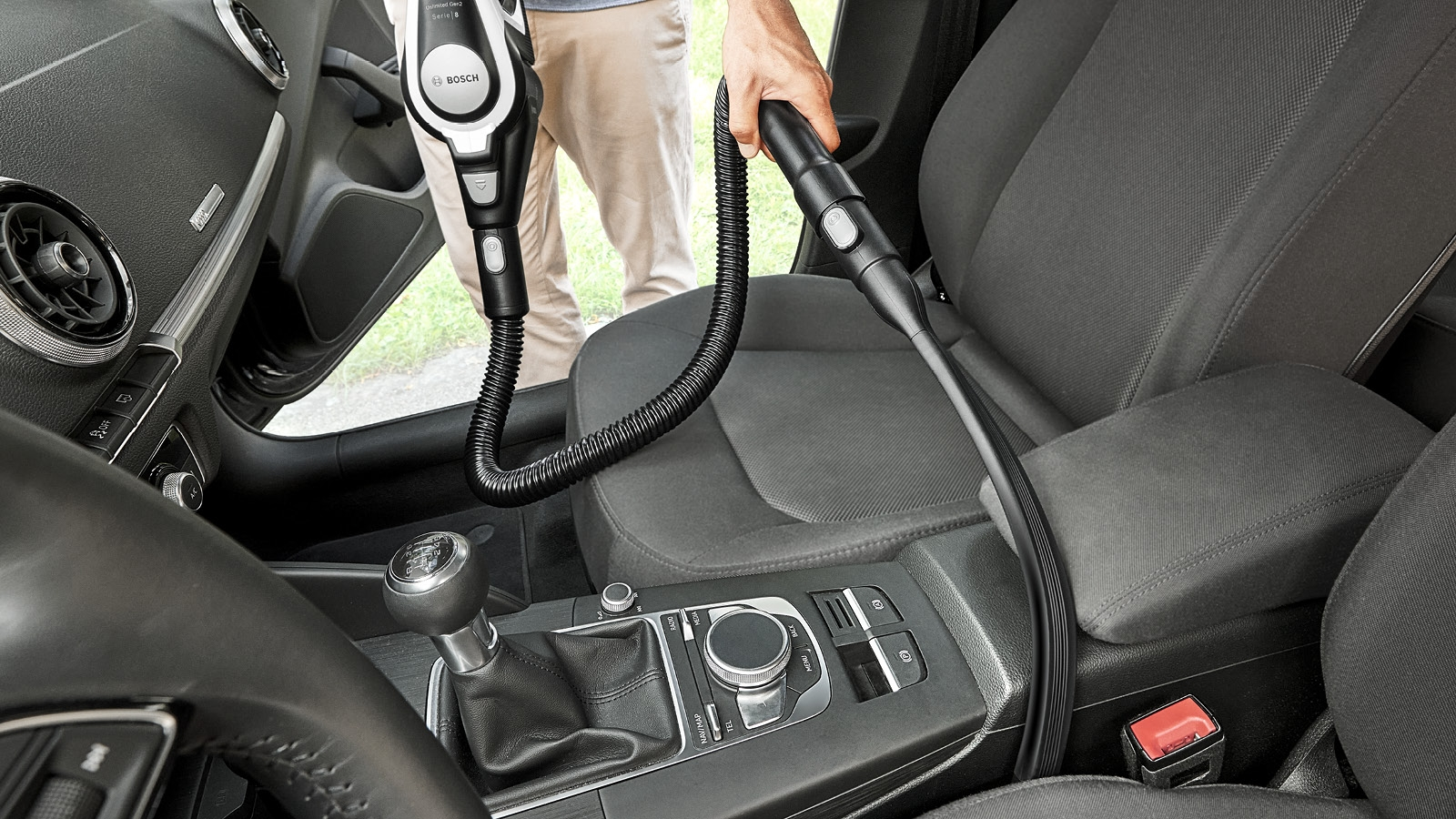 Eine Person saugt einen Autoinnenraum. Ein Bosch-Staubsauger wird auf einem grauen Sitz verwendet. Das Armaturenbrett und der Schalthebel des Autos sind sichtbar.
