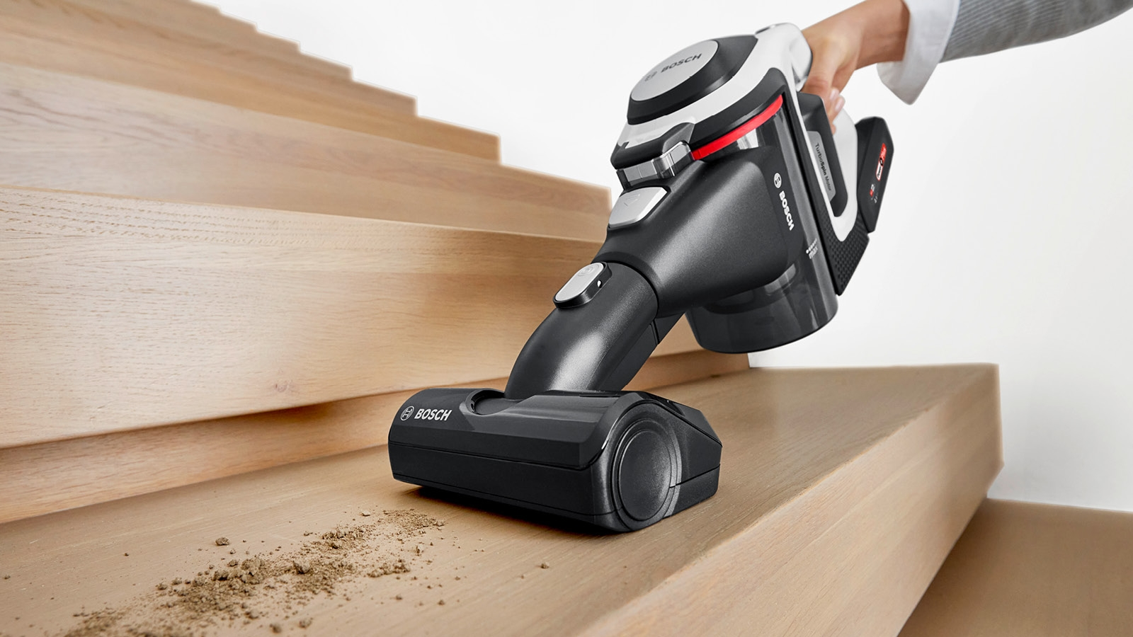 Person reinigt Holztreppe mit kabellosem Bosch-Staubsauger. Der Staubsauger ist schwarz-weiß und entfernt Schmutz.