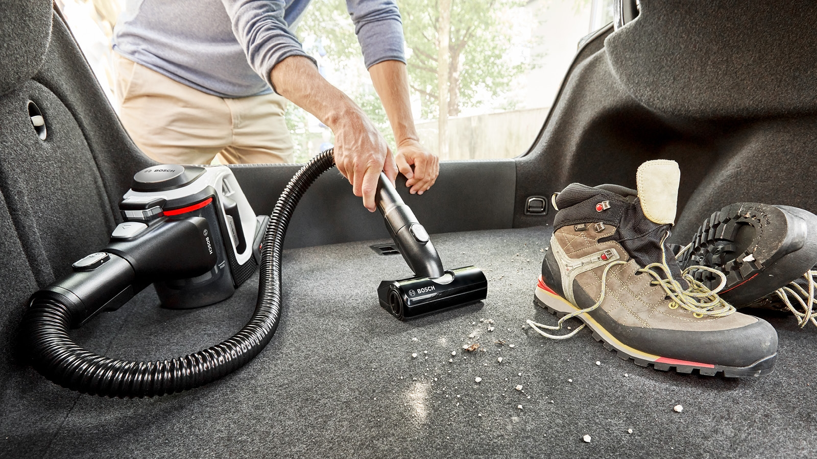 Eine Person saugt den Kofferraum eines Autos. Ein Staubsauger wird in der Nähe von Wanderstiefeln verwendet. Der Kofferraum ist mit Teppich ausgelegt.