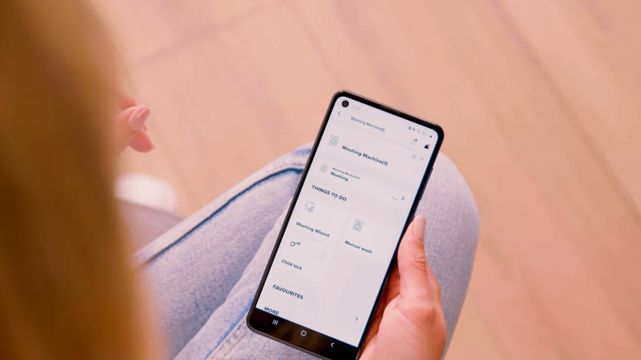Osoba trzymająca smartfona wyświetlającego interfejs aplikacji pralki.